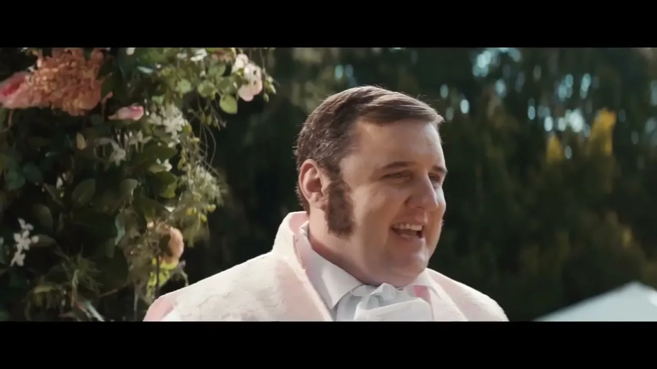 A man in a pink period costume smiles and looks off-camera, standing outdoors next to a floral arrangement.
