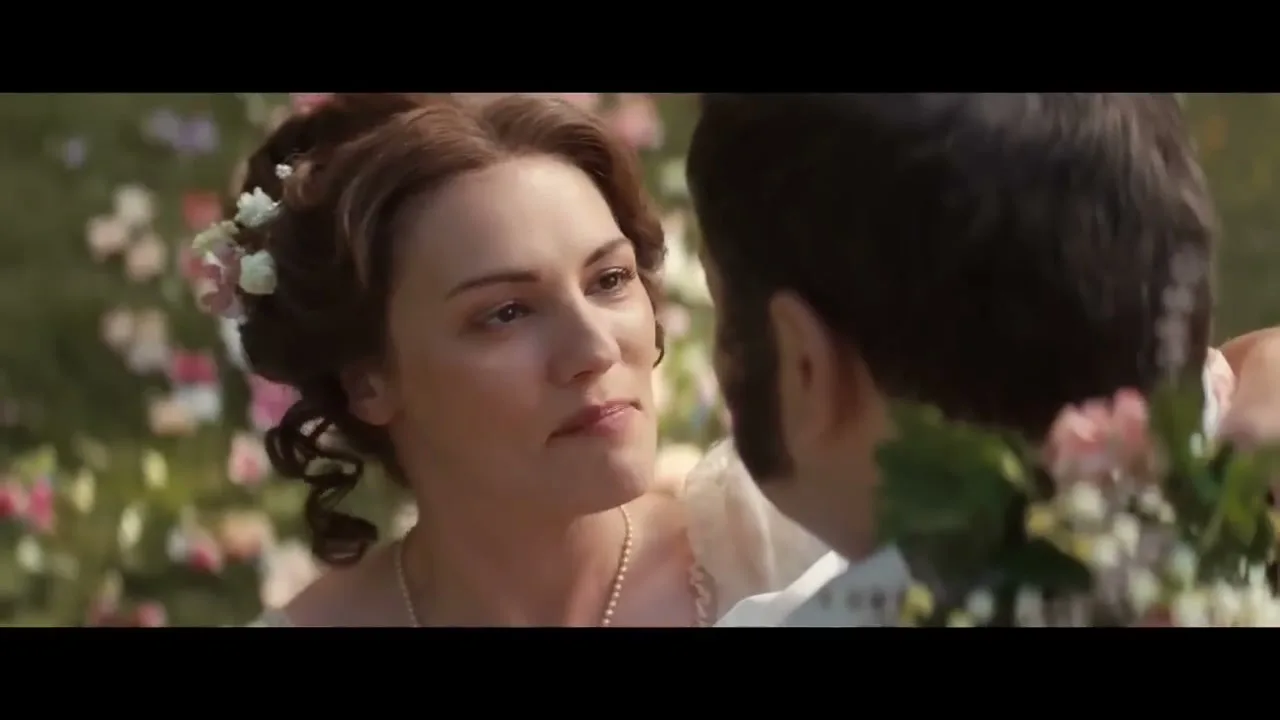 A woman with flowers in her hair, wearing a pearl necklace and a white dress, smiles and talks to a man in a garden.
