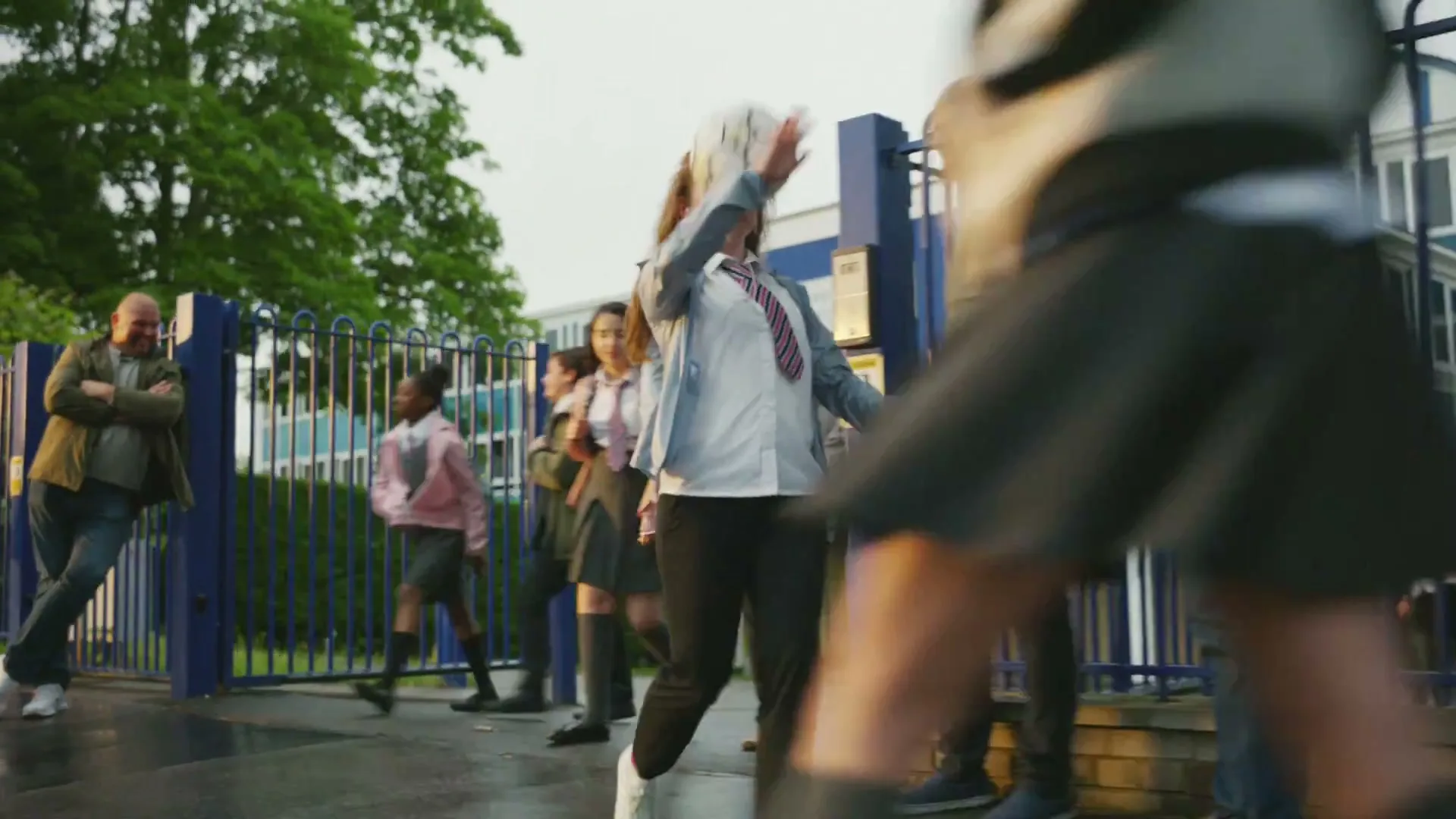 A young female student in a school uniform juggles a football with her head and feet outside a school building, as other students and adults walk by.