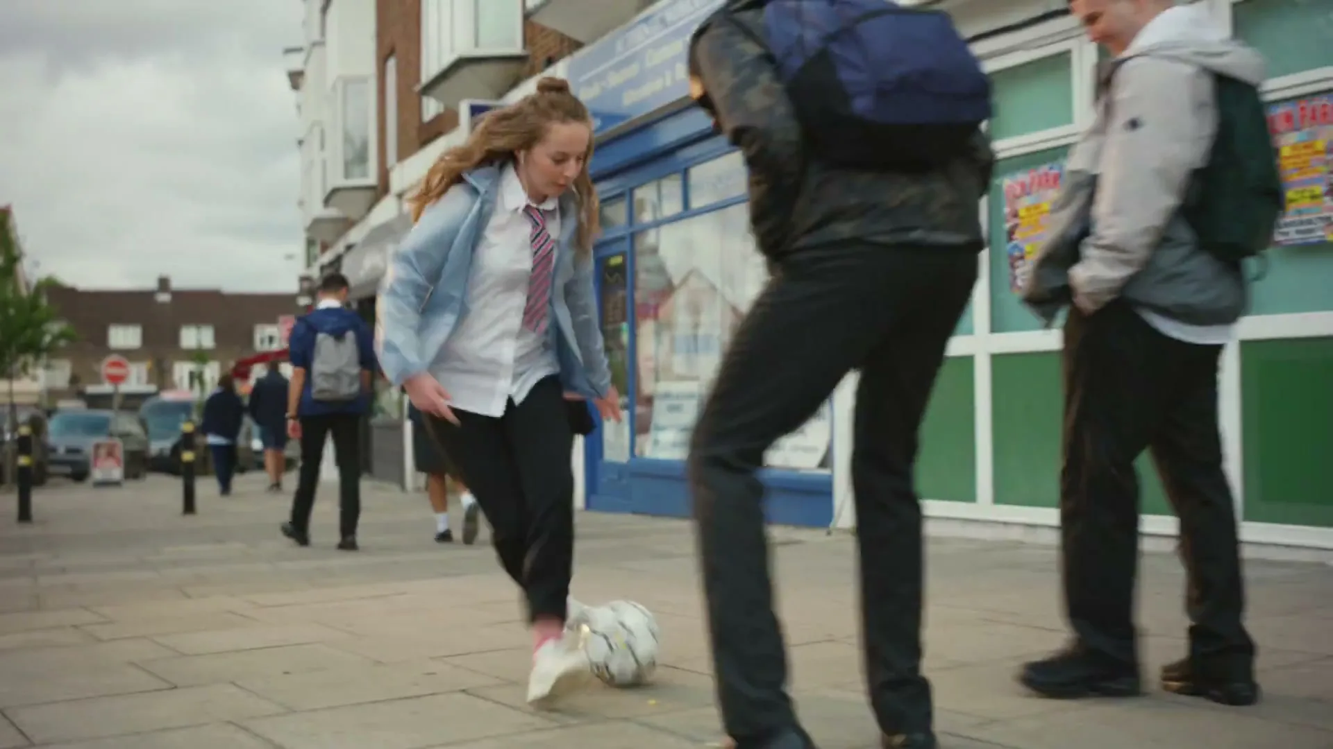 A female teen in school uniform dribbles a football on a busy urban sidewalk, passing other students and storefronts under an overcast sky.