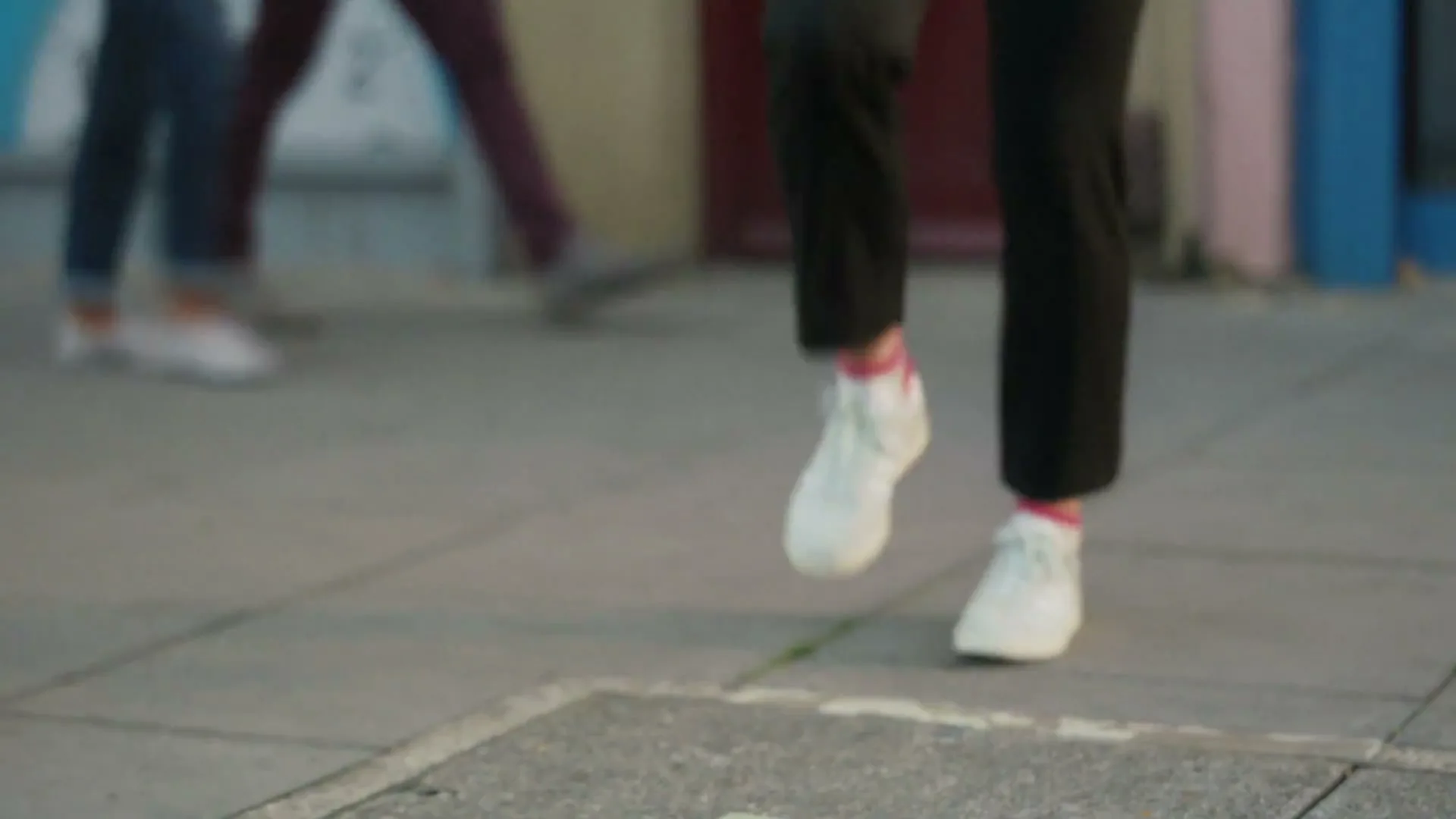 A person in black pants and white sneakers juggles a soccer ball on a paved surface, with other people in the background.