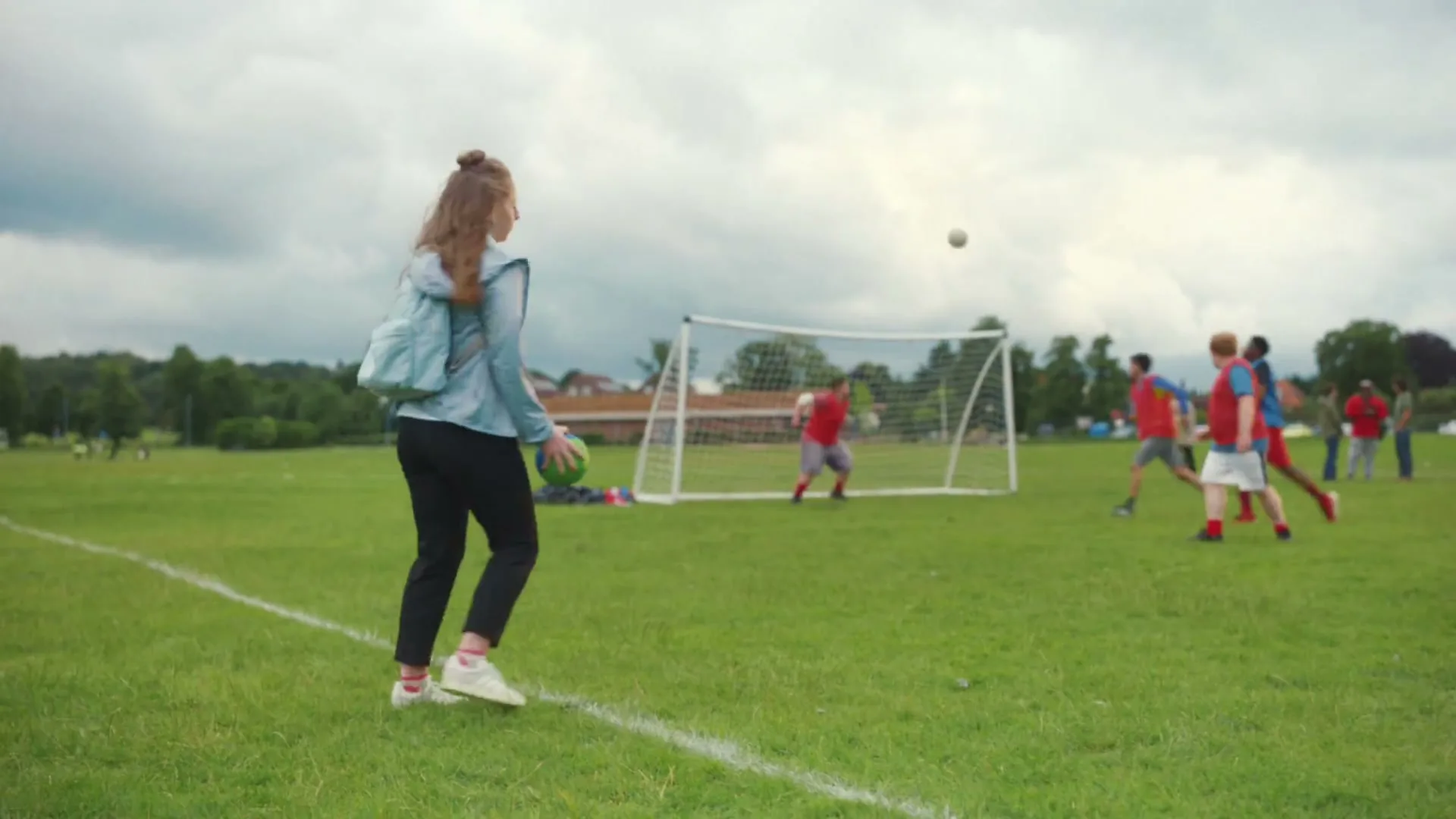 A young woman in a school uniform and light blue jacket juggles a soccer ball on a grassy field, while other people play soccer in the background.