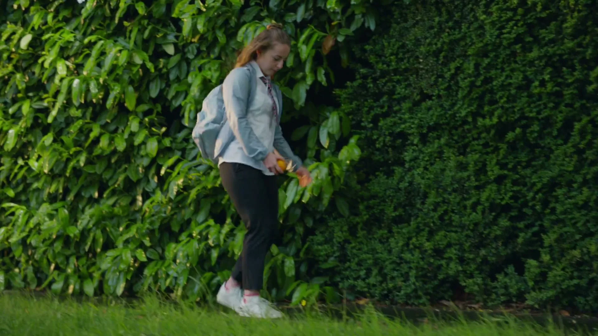 A young female student in school uniform struggles to put on a backpack and drops an orange on the grass next to a hedge.