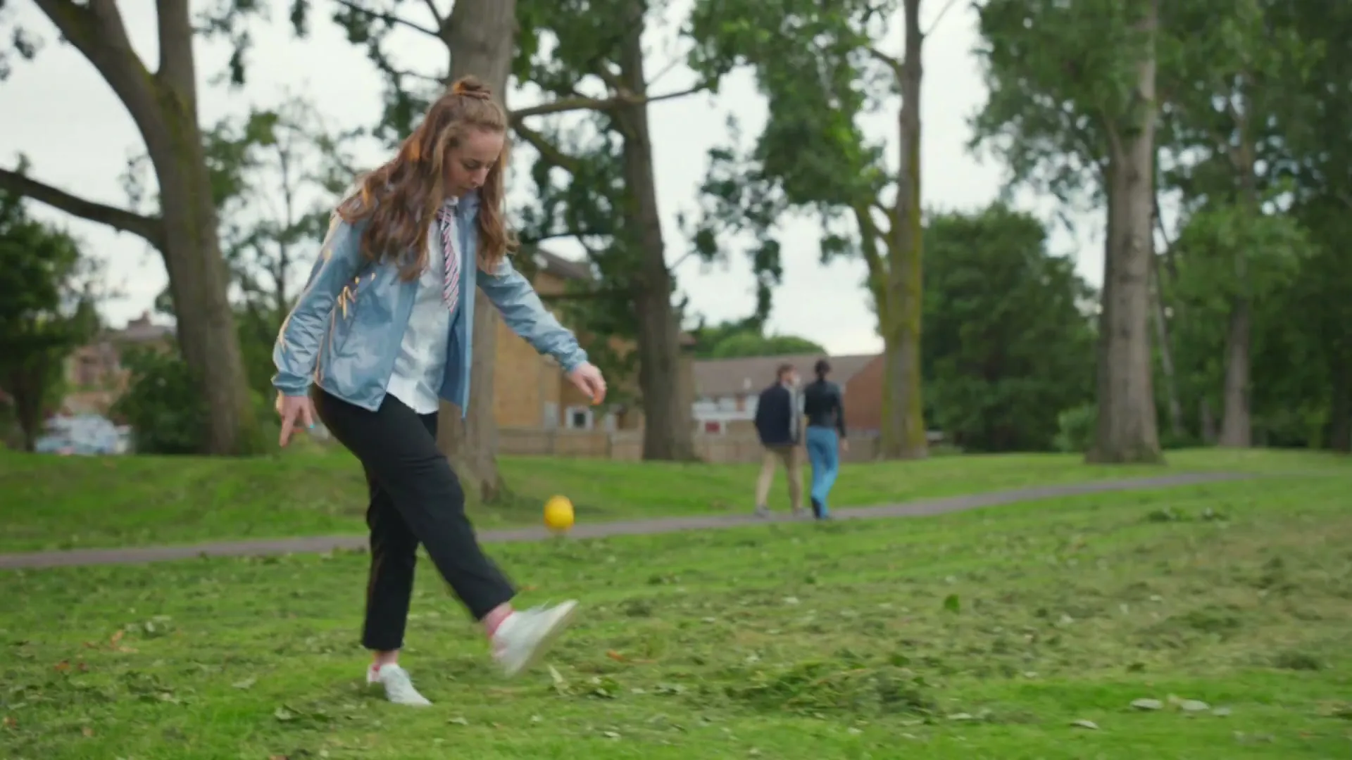 A young female student in a school uniform and light blue jacket practices kicking a small yellow ball in a grassy park.