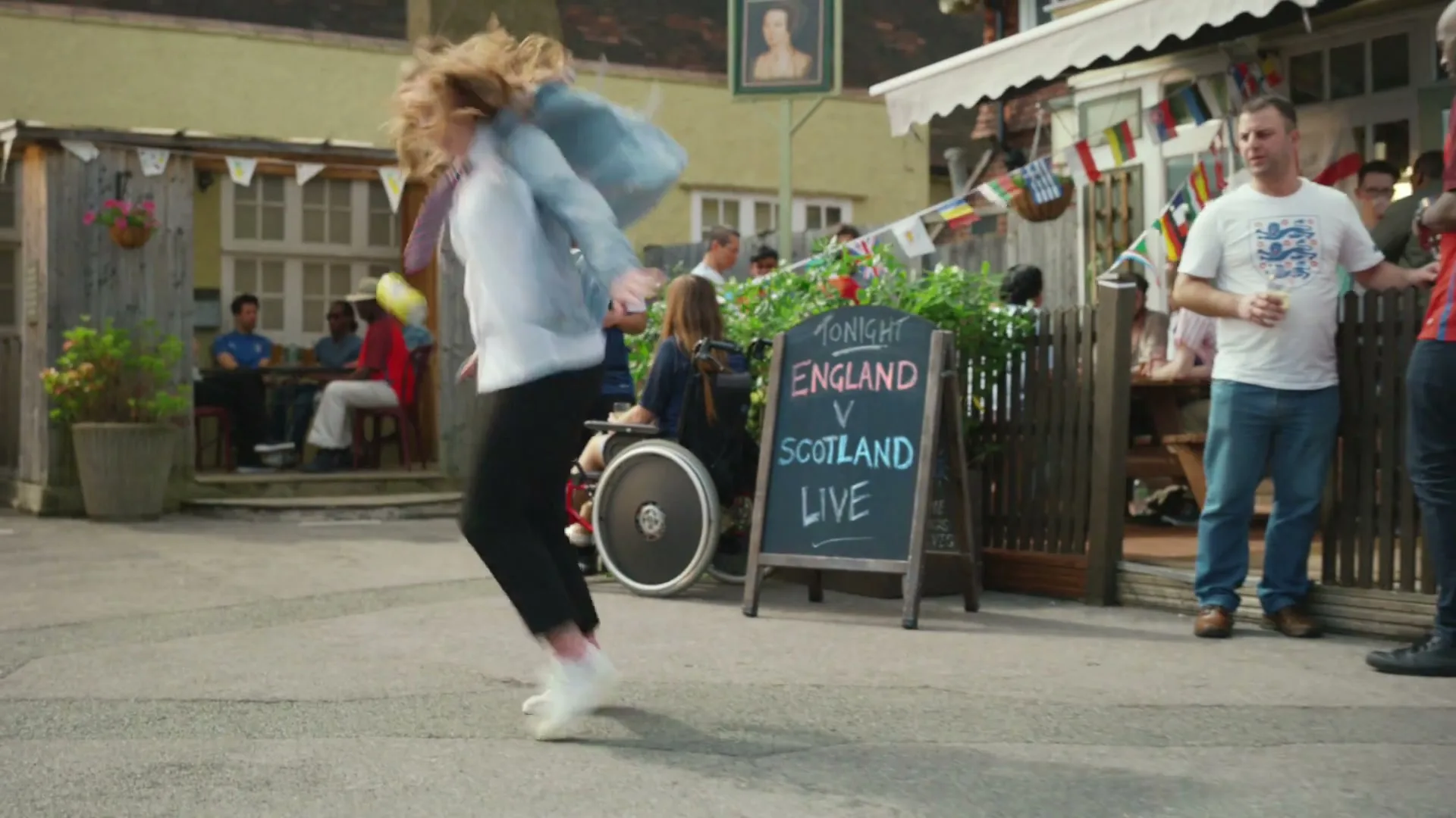 A young woman in a denim jacket jumps over a small yellow can in a pub garden, while other patrons watch and a chalkboard advertises a football match.