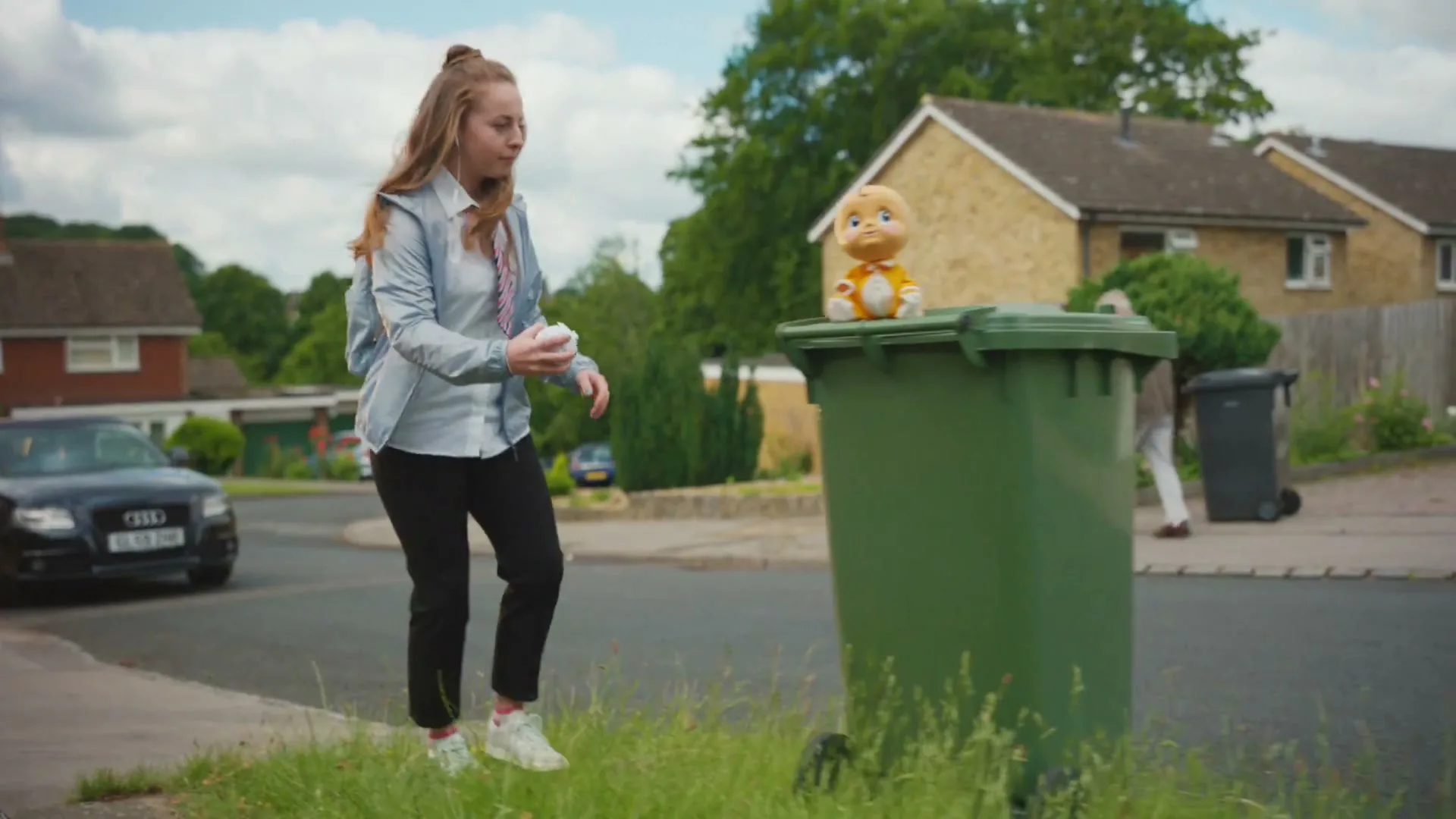 A young woman in a school uniform kicks a crumpled paper ball towards a trash can with a doll on top, on a suburban street.