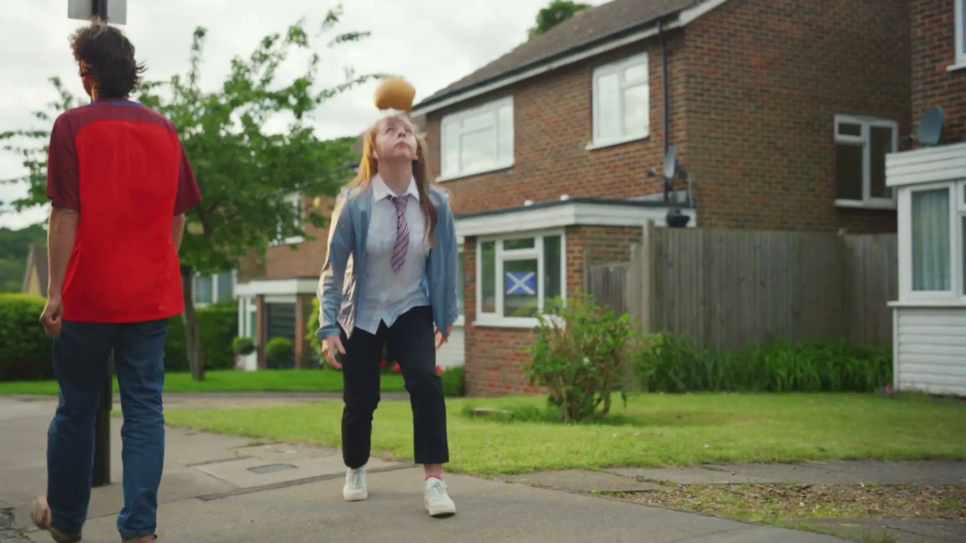 A young girl in a school uniform practices keepy-uppies with a football on a suburban sidewalk as a man walks past her.