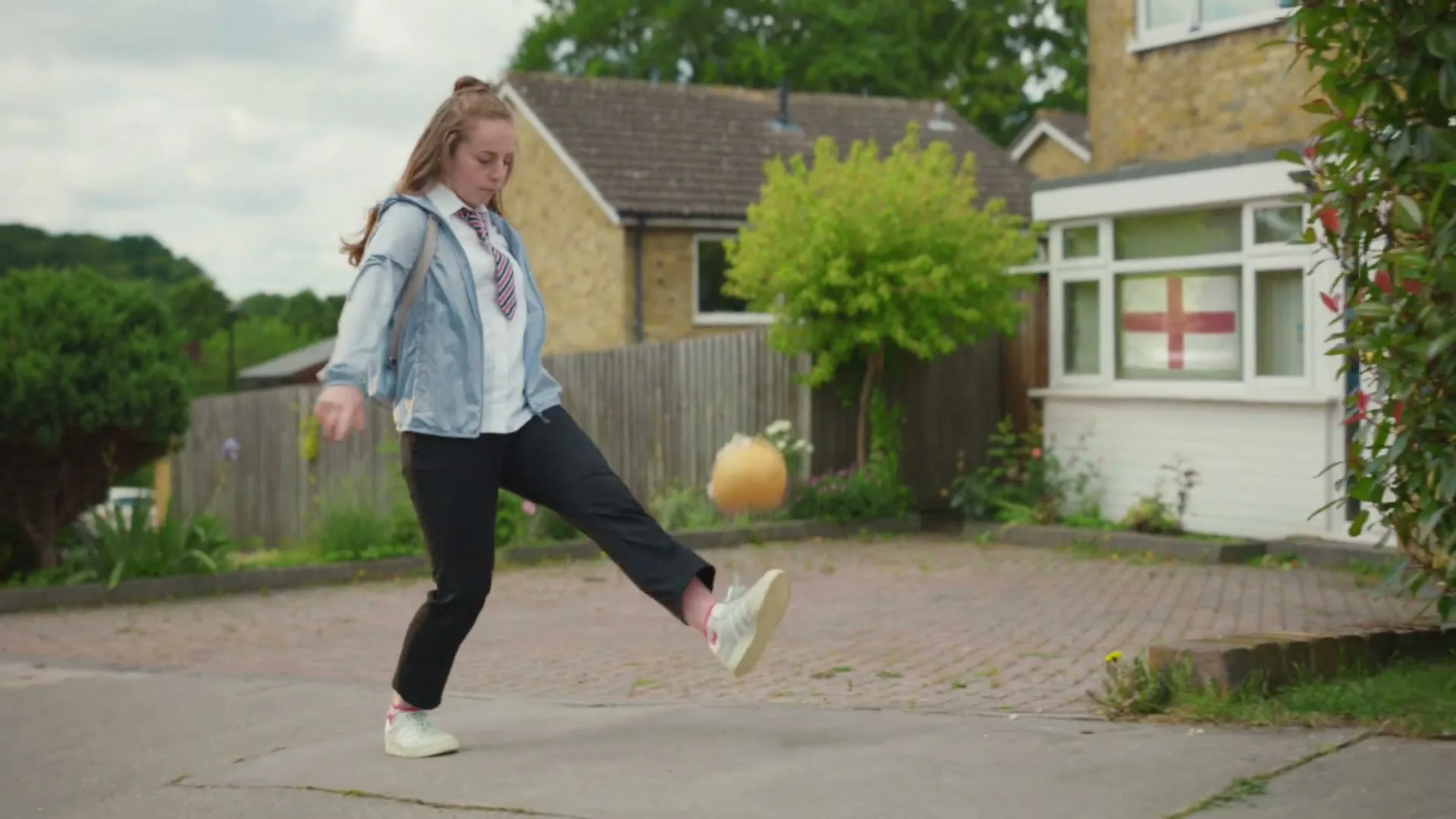 A young female student in school uniform and a light blue jacket juggles a football in a suburban driveway with a house displaying an England flag in the background.
