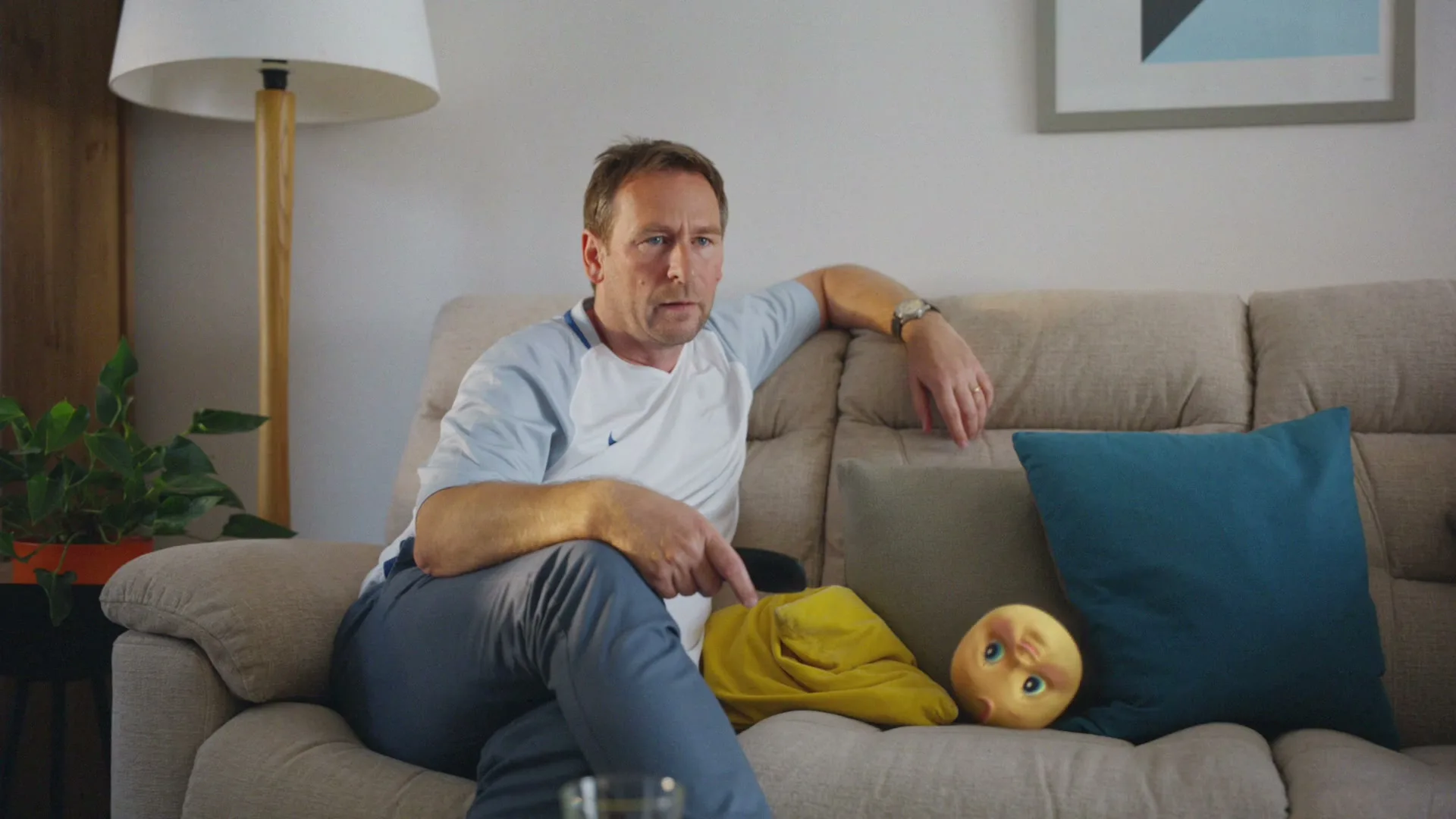 A middle-aged man in a t-shirt and sweatpants sits on a sofa, looking intently at something off-screen, with a doll's head next to him.