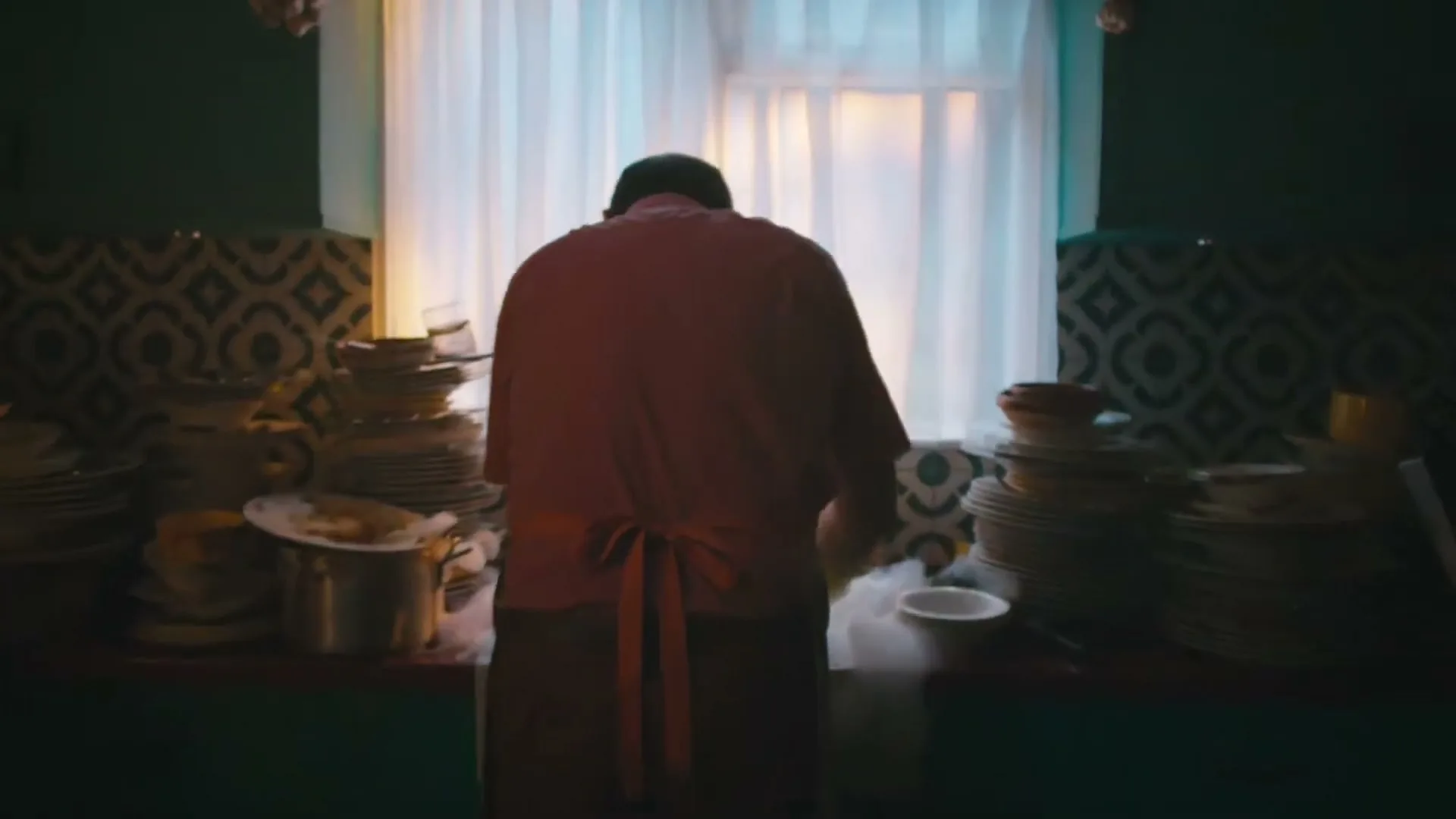 A man in a pink shirt and apron washes a huge pile of dishes in a dimly lit kitchen, then turns to the camera with a creepy smile.