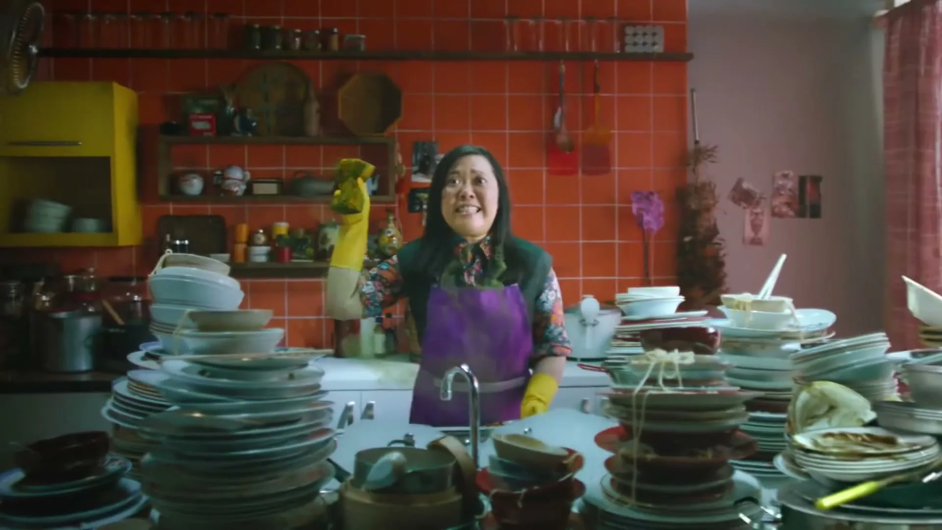 A woman in a green dress washes dishes in a sink, then another woman in an apron and yellow gloves holds up a dirty sponge and smiles maniacally in a kitchen.
