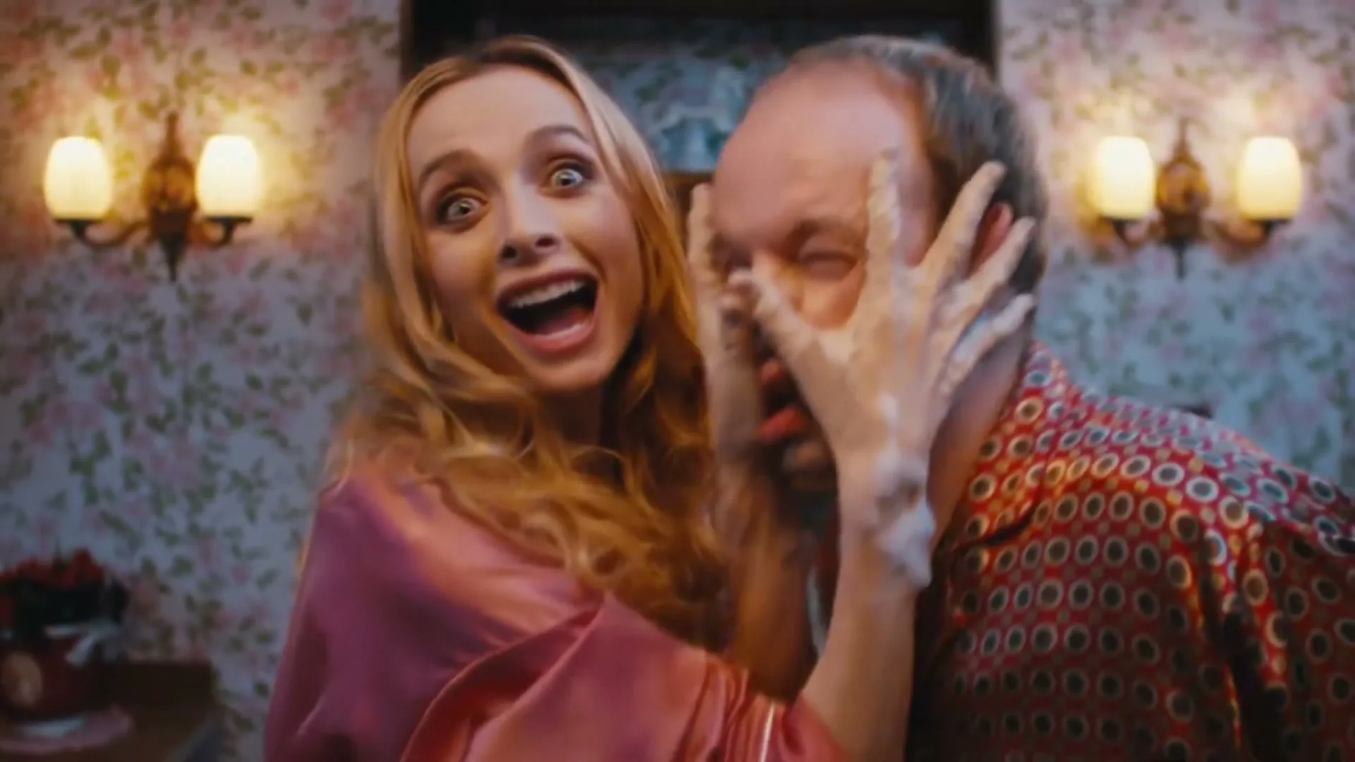 A woman with wild eyes and a wide smile, wearing a pink robe, embraces a man from behind, covering his face with her hands in a kitchen.