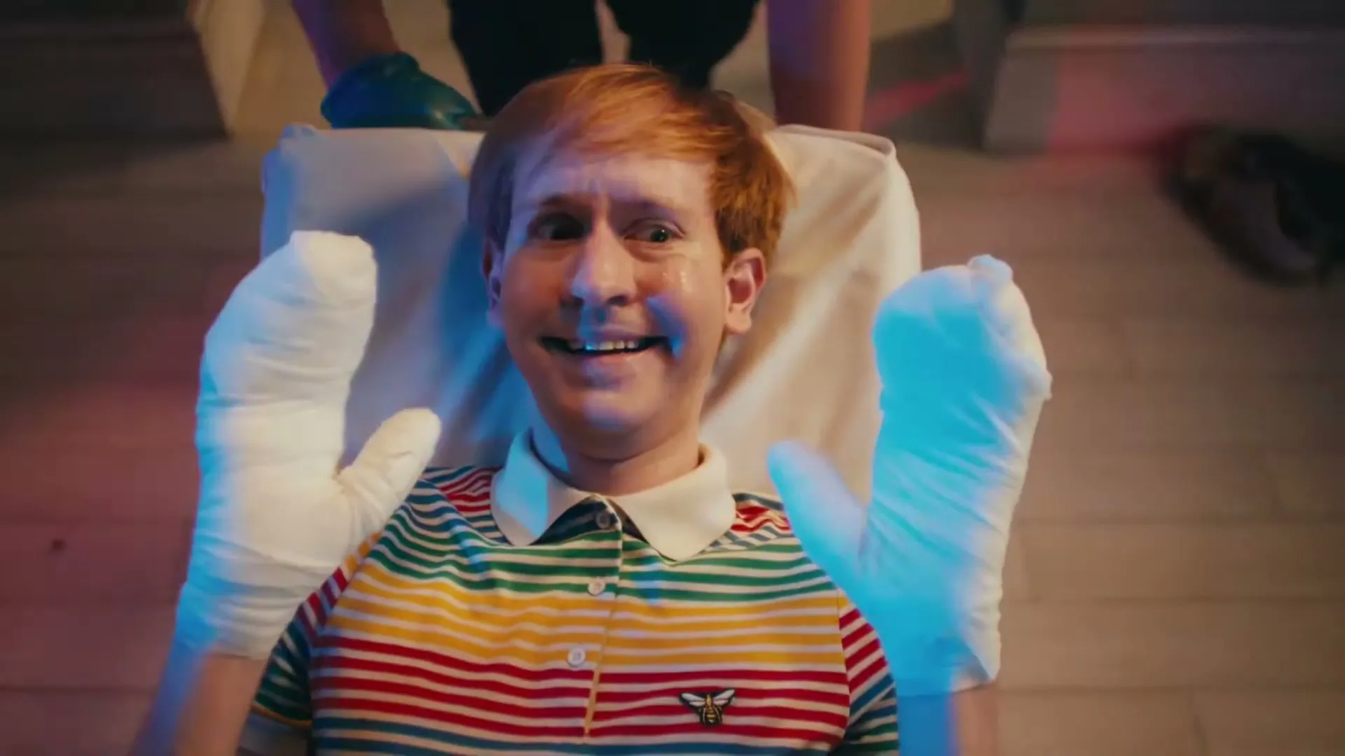 A man with red hair and a striped shirt lies on a table, smiling maniacally with his hands wrapped in white bandages, as someone in blue gloves holds his arms.
