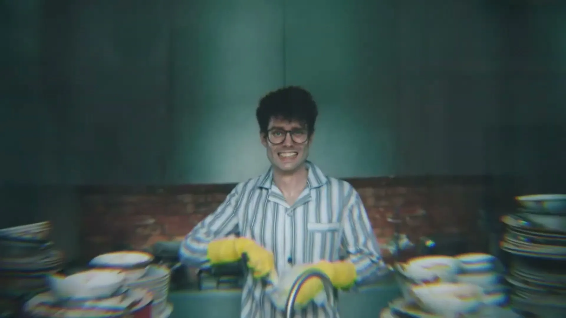 A man in pajamas and yellow gloves washes dishes in a messy kitchen, looking distressed as the camera pulls back to reveal him through a window.