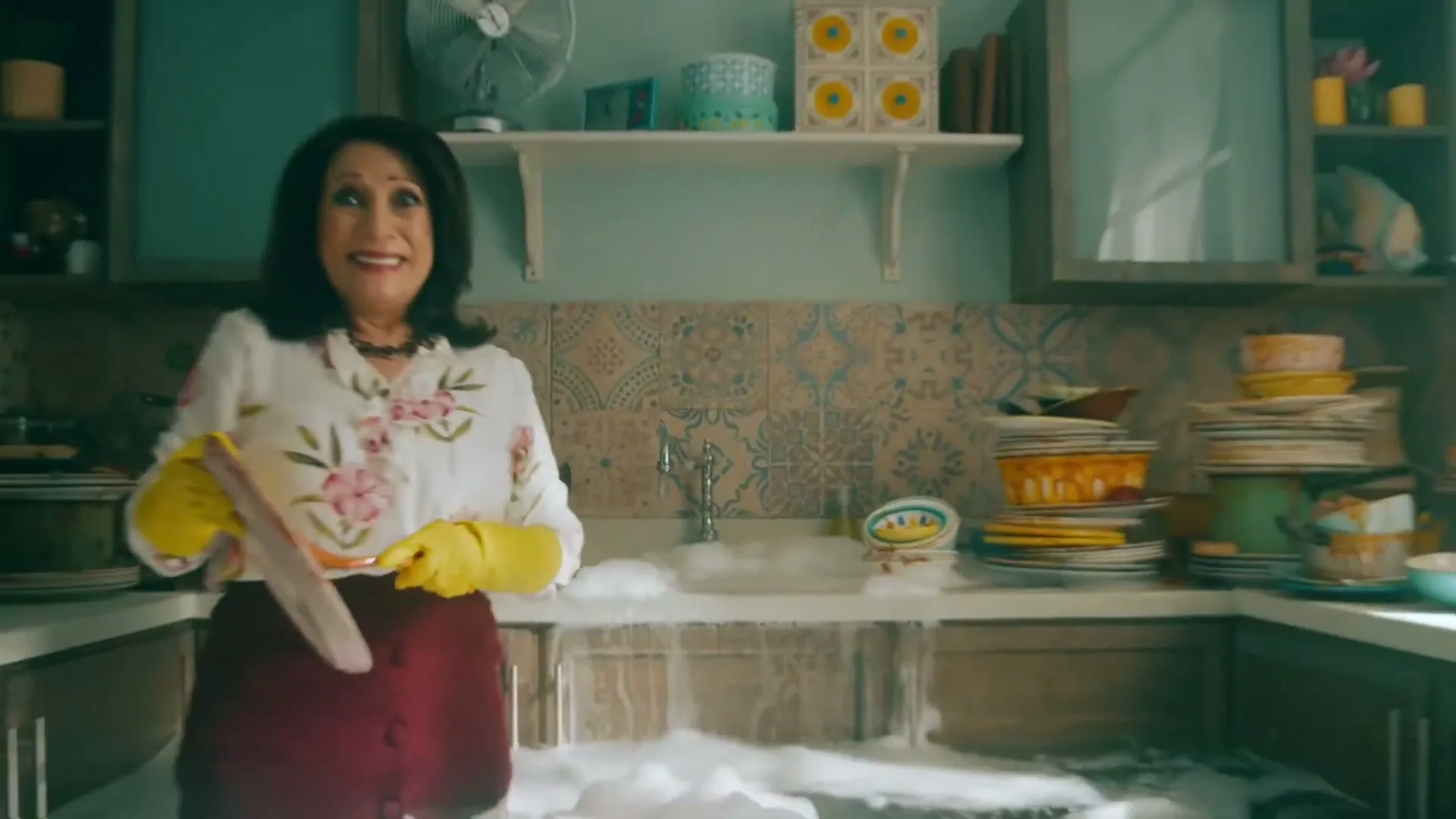 A woman in a floral shirt and yellow gloves stands in a kitchen, smiling as water and bubbles overflow from the sink onto the floor.