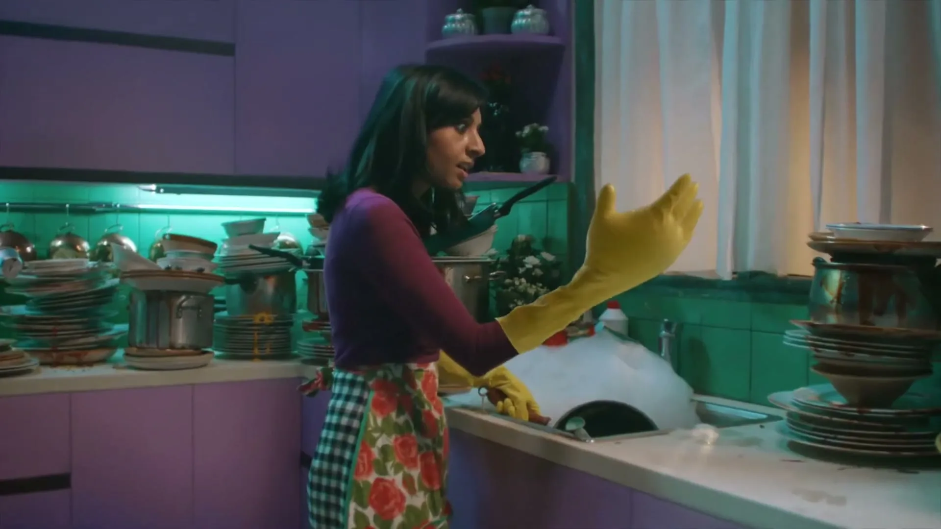 A woman in a purple shirt and floral apron, wearing yellow dish gloves, smiles at the camera while washing dishes in a brightly lit kitchen.