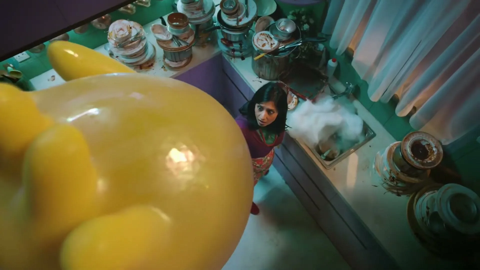 A woman in a messy kitchen looks up in fear as a giant yellow rubber glove descends from above, filling the frame.