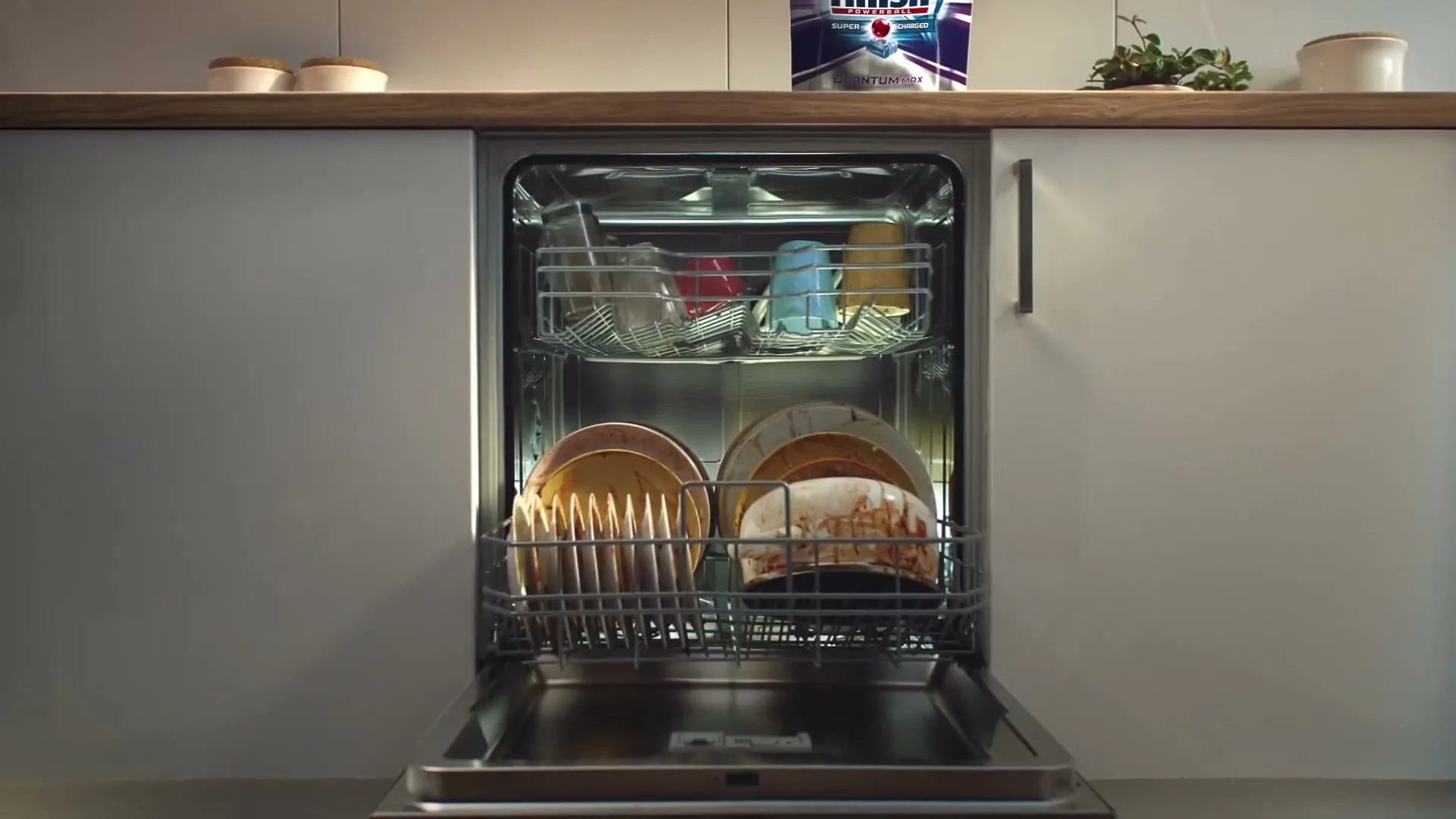 A person loads dirty dishes into an open dishwasher in a modern kitchen, with a Finish product visible on the counter.