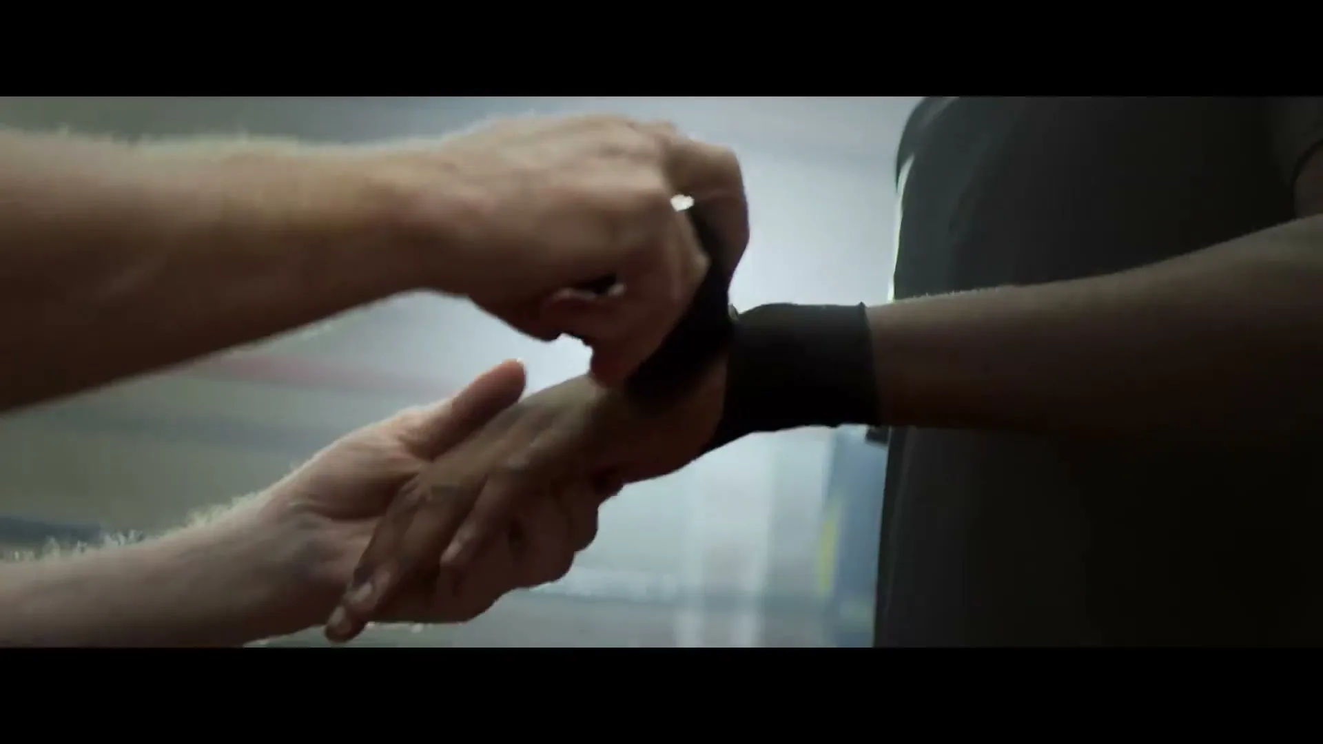 A close-up shot shows a white man's hands wrapping a black hand wrap around a Black man's wrist in a dimly lit boxing gym.