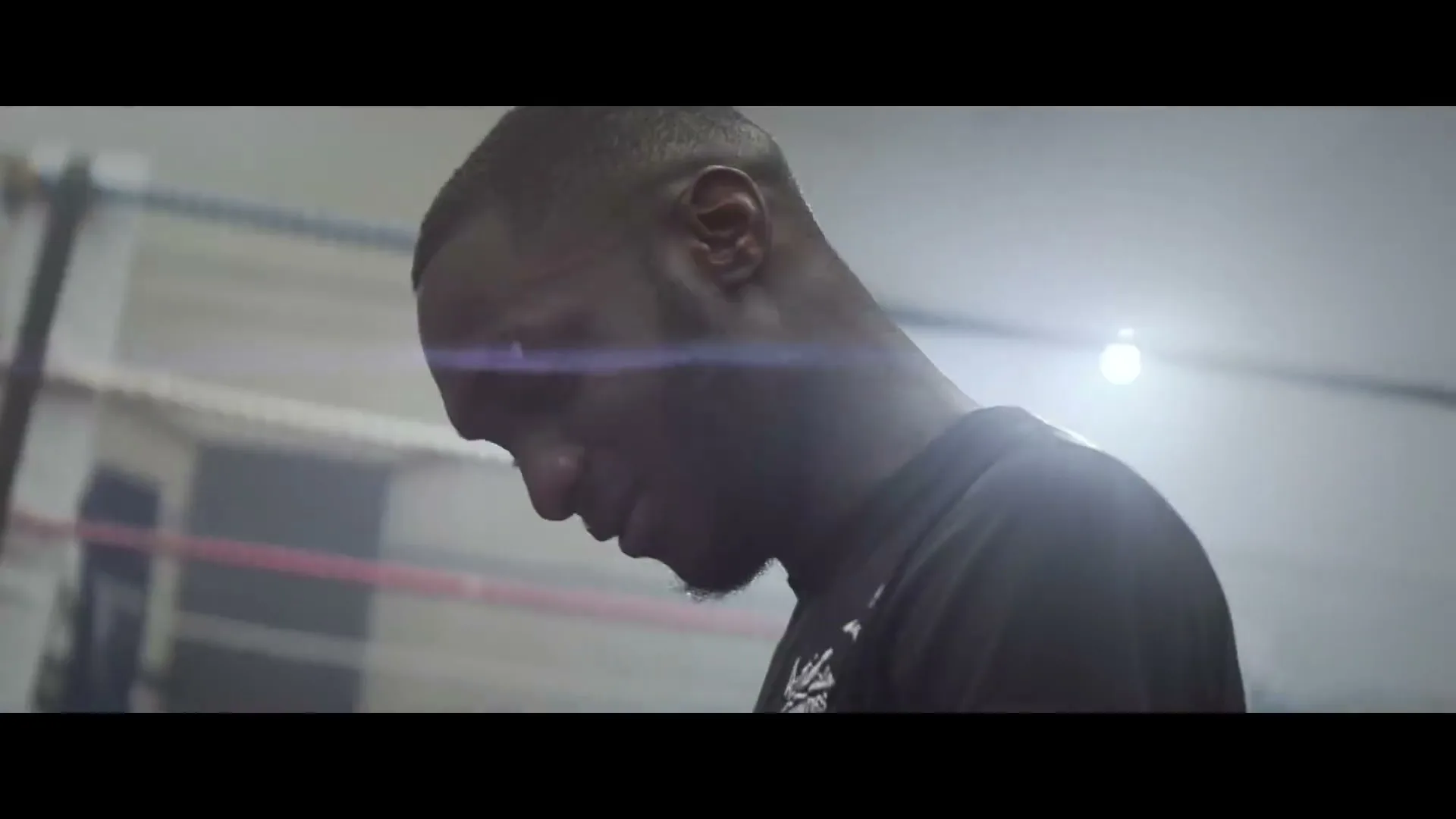 A young Black man with a fade haircut and beard, wearing a black t-shirt, looks down in a boxing ring, appearing to be in deep thought.