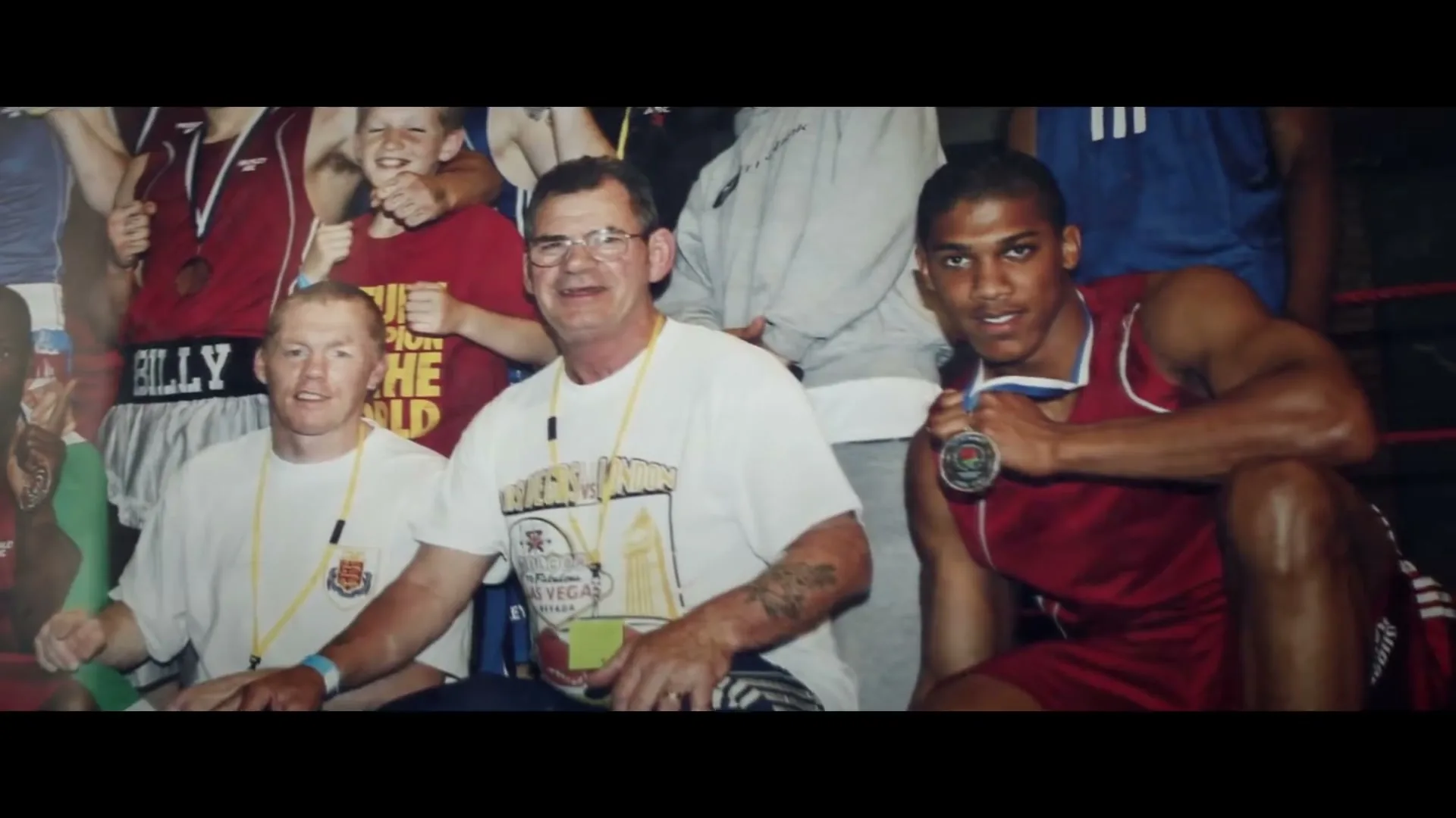 A young Anthony Joshua, wearing a red boxing uniform and a medal, poses with a group of men and boys in a boxing ring.