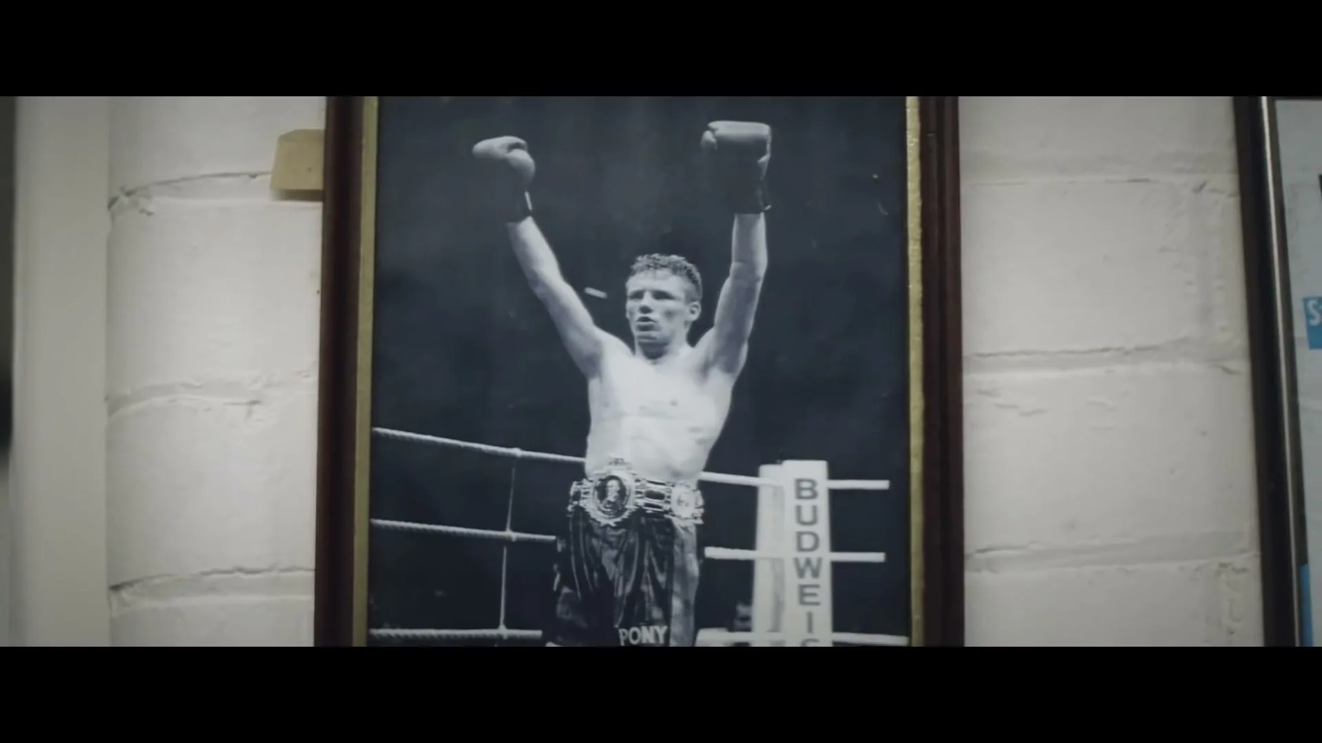 A black and white framed photo of a young male boxer with his arms raised in victory, wearing boxing gloves and a championship belt, hangs on a white brick wall.