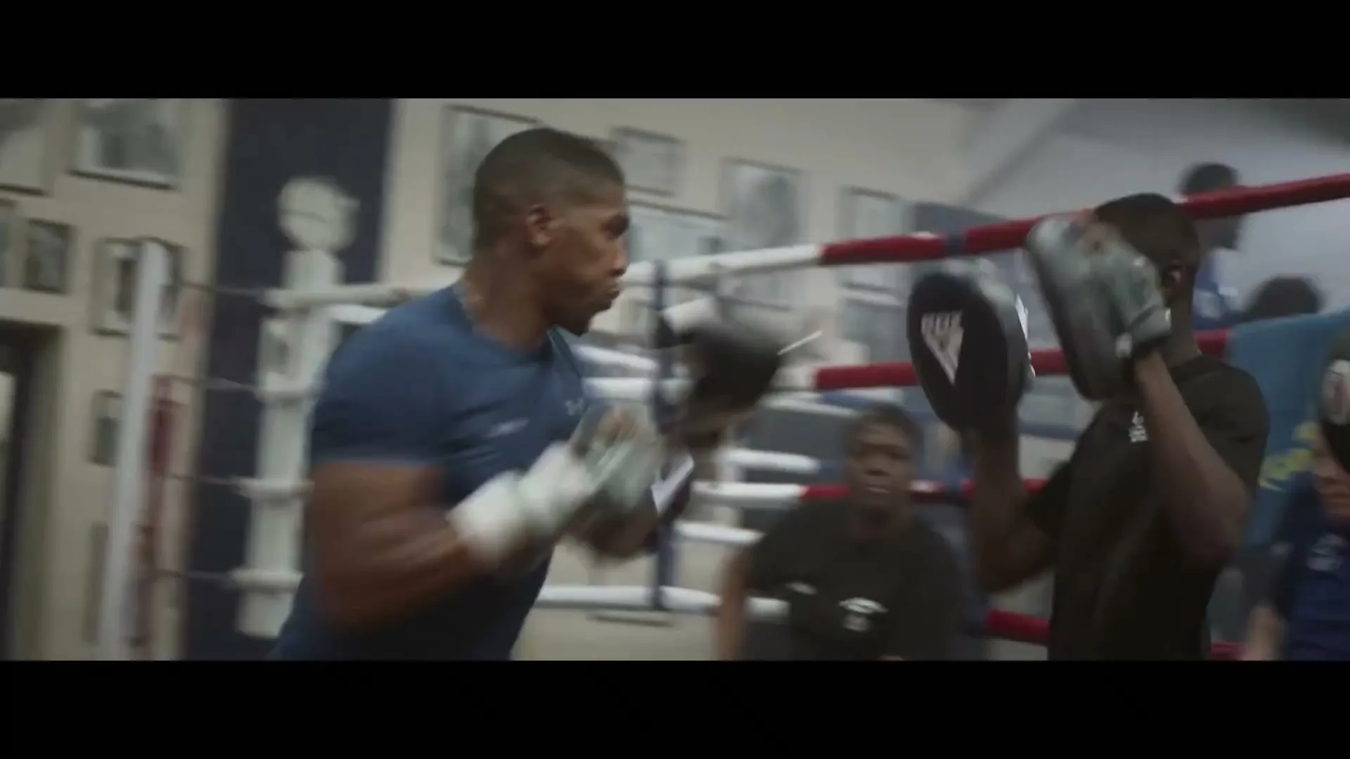 A male boxer in a blue shirt and black gloves trains with a partner holding pads in a boxing gym.