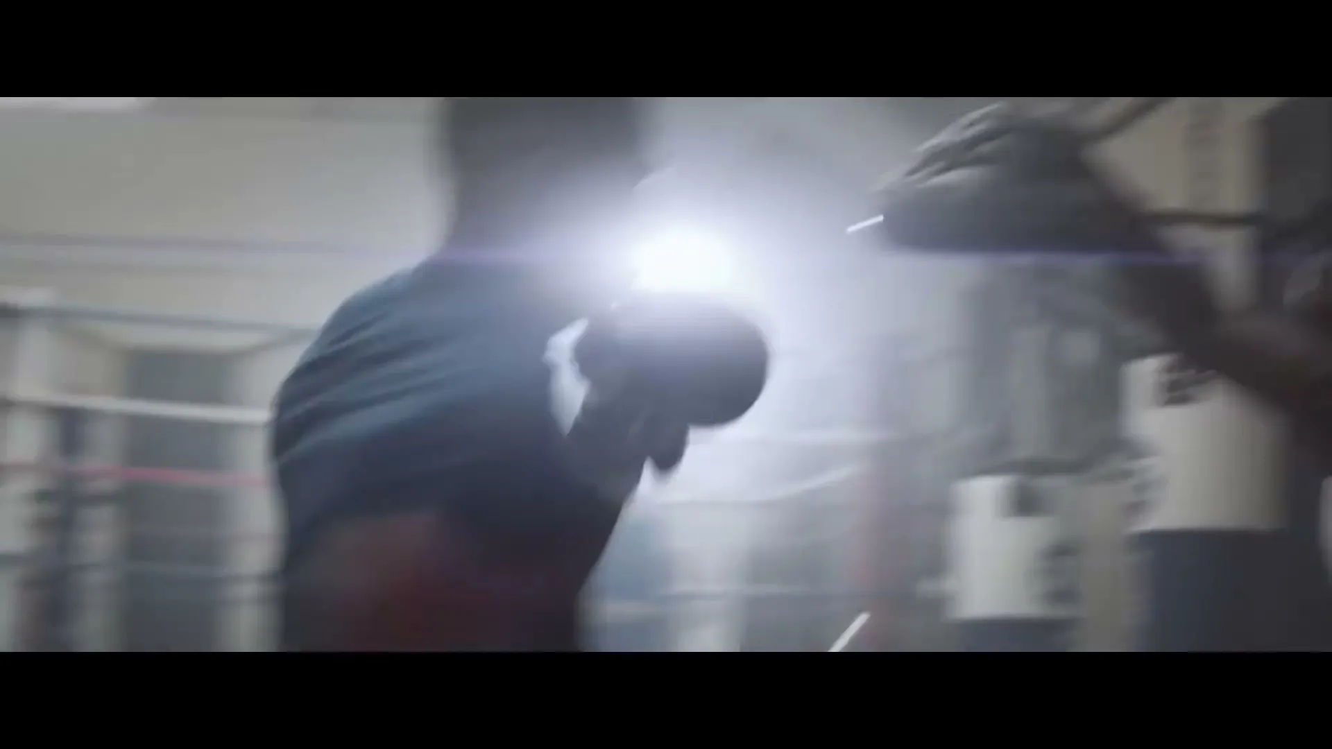 A male boxer in a blue shirt trains with a coach, punching pads in a dimly lit boxing gym with a bright light flare.