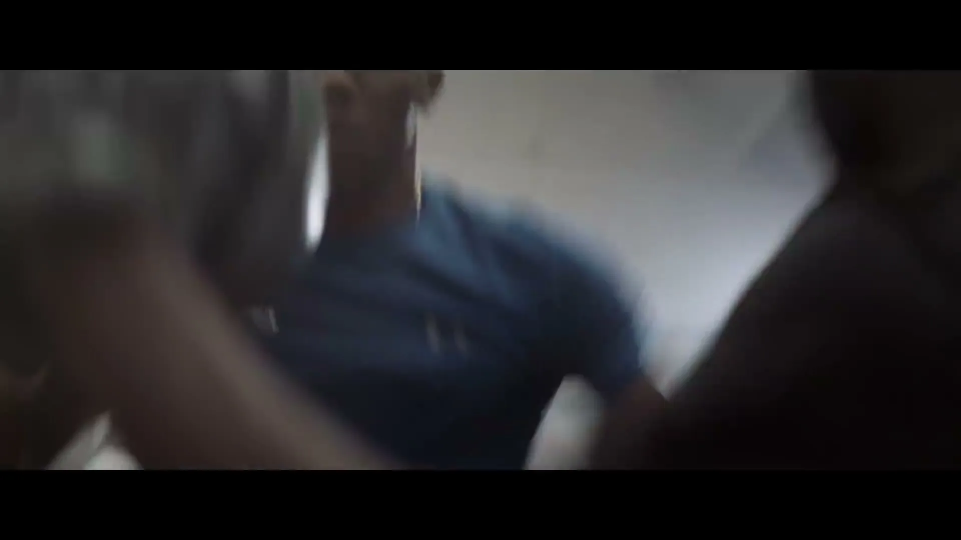 A male boxer in a blue shirt is seen from a low angle, training with a punching bag in a gym.