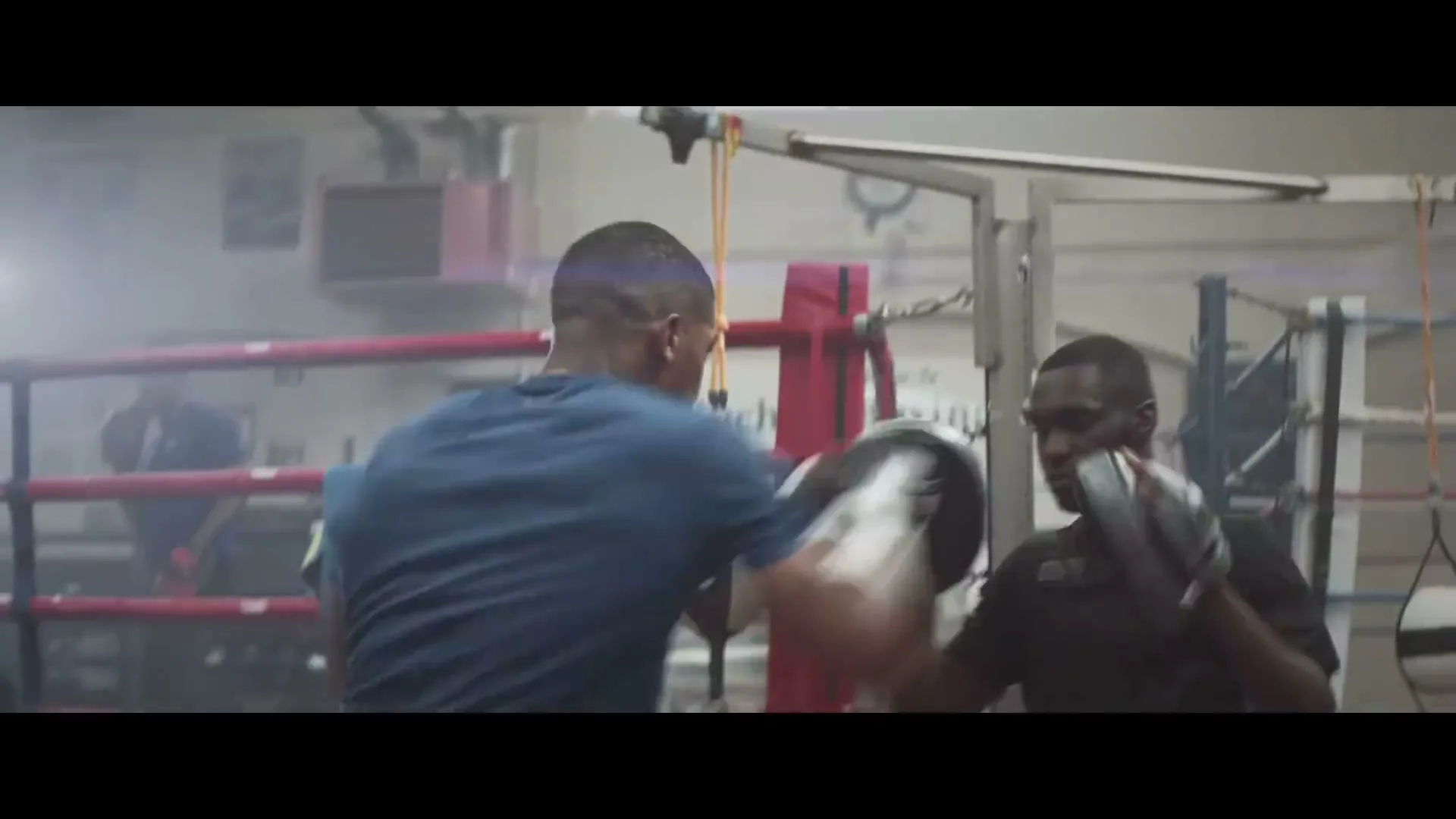 A male boxer in a blue shirt trains with a coach in a boxing ring, throwing punches at mitts in a gym.