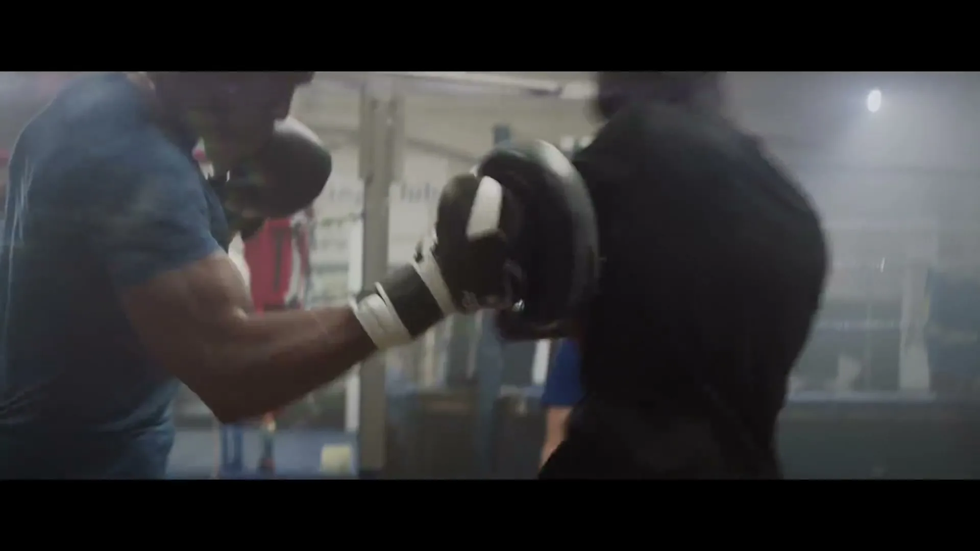 Two male boxers, one wearing a blue t-shirt and boxing gloves, punches pads held by another man in a boxing gym.