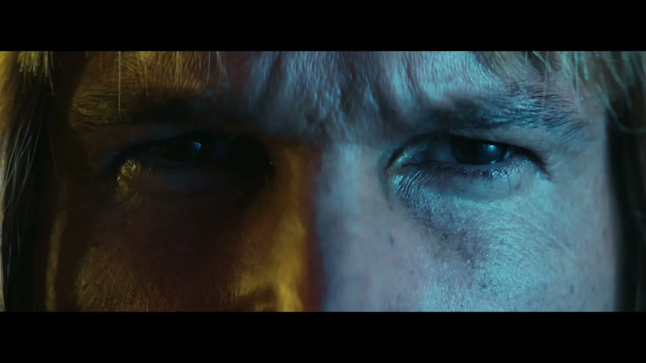 A man's eyes are shown in an extreme closeup, with half his face lit by warm light and the other half by cool blue light, as he looks around.