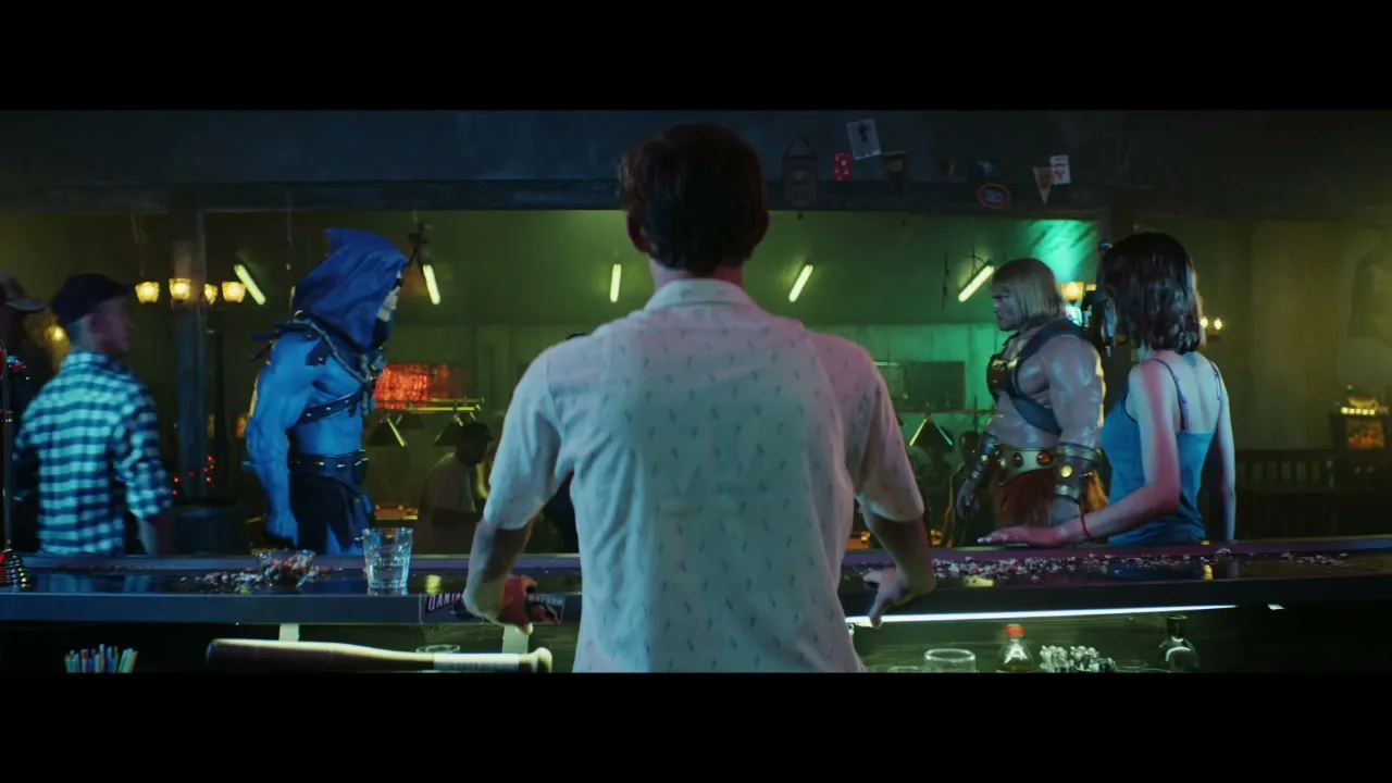 A man in a white shirt stands at a bar, looking at Skeletor and He-Man who appear on either side of him, in a dimly lit bar.