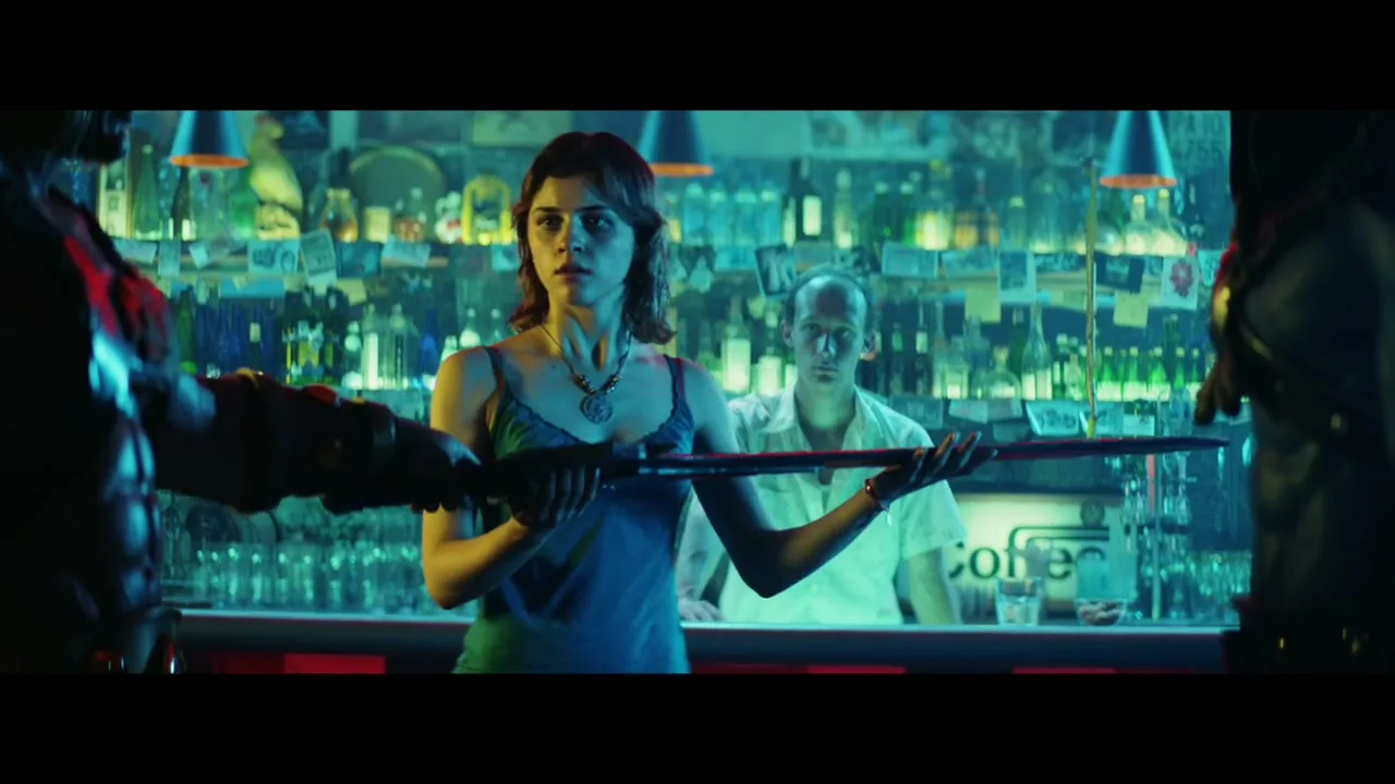 A young woman in a blue top stands at a bar, looking intently at something off-screen, while a bartender watches from behind her.