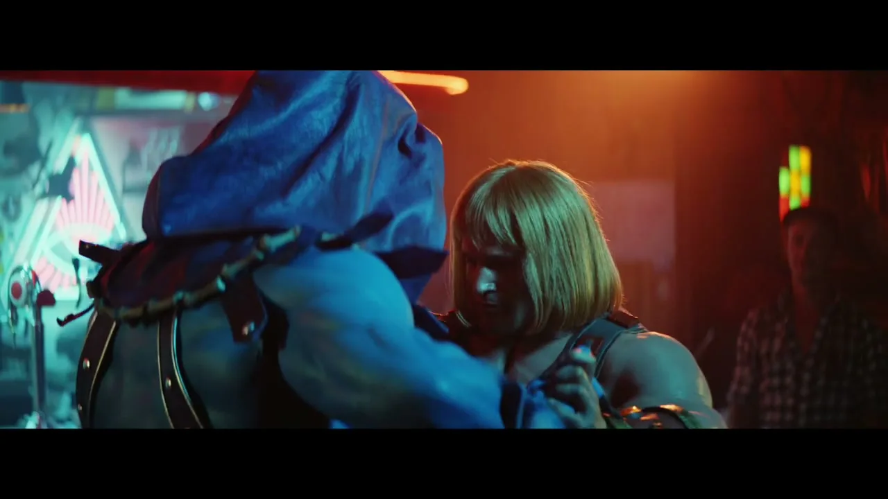He-Man, a muscular man with blonde hair, dances closely with Skeletor, a blue-skinned figure, in a dimly lit bar.
