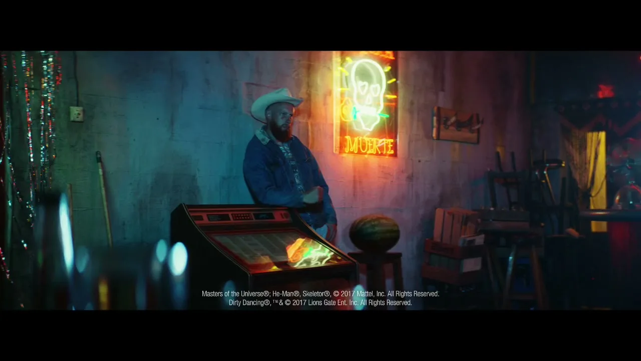 A man in a cowboy hat and denim jacket leans against a wall next to a jukebox, under a neon skull sign that says "MUERTE" in a dimly lit bar.
