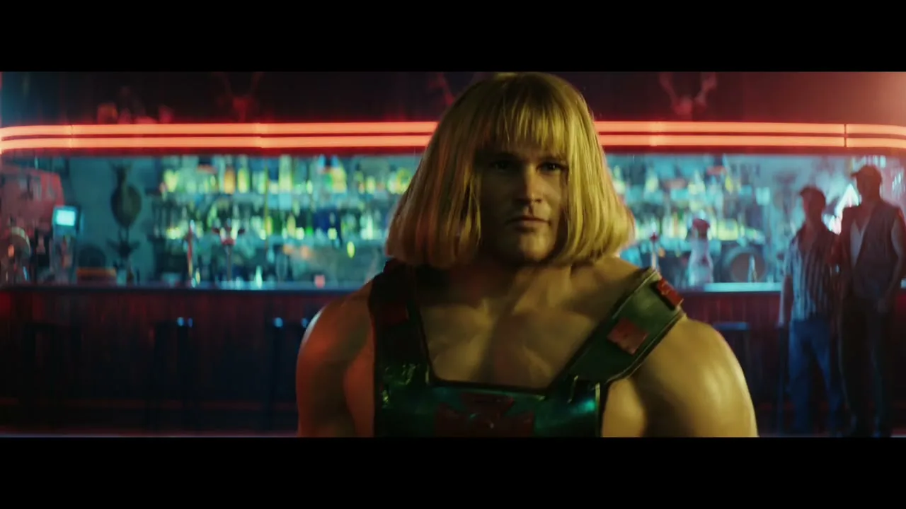 He-Man, a muscular man with blonde hair and green and red straps, turns around in a bar with Skeletor in the background.