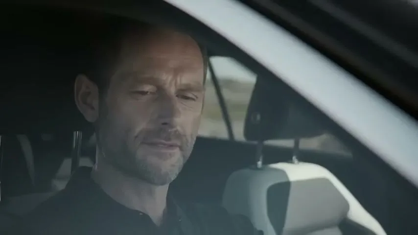 A bald man with a beard sits in the driver's seat of a car, looking down and to the side, in a medium closeup shot.