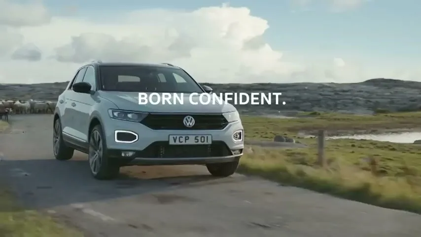 A white Volkswagen T-Roc drives on a winding road through a rural landscape with sheep in the background under a bright sky.