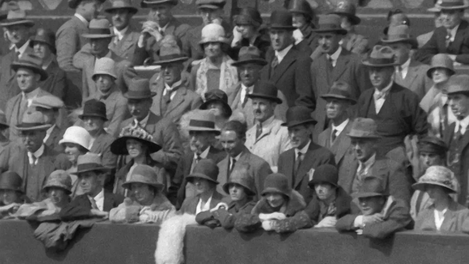 A crowd of men and women in formal attire and hats watch a tennis match from the stands at Wimbledon in black and white archival footage.