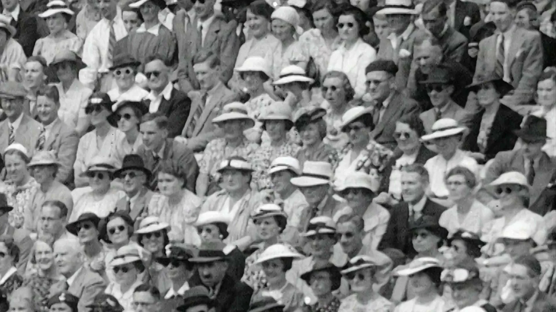 A black and white archival shot shows a crowd of people in formal attire watching a tennis match at Wimbledon.
