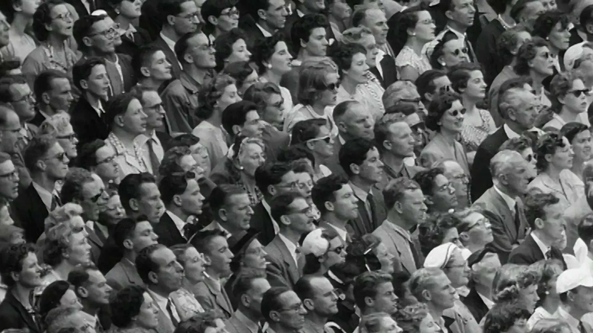 A black and white shot of a large crowd of people, mostly adults, watching an event with varied expressions in an outdoor stadium.