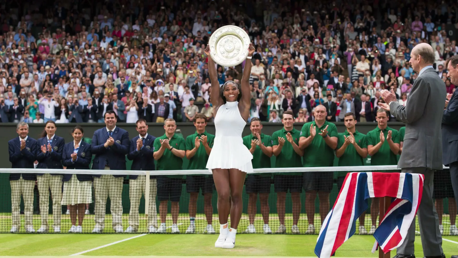 Serena Williams, a female tennis player in a white dress, raises a trophy on a tennis court to a cheering crowd at Wimbledon.