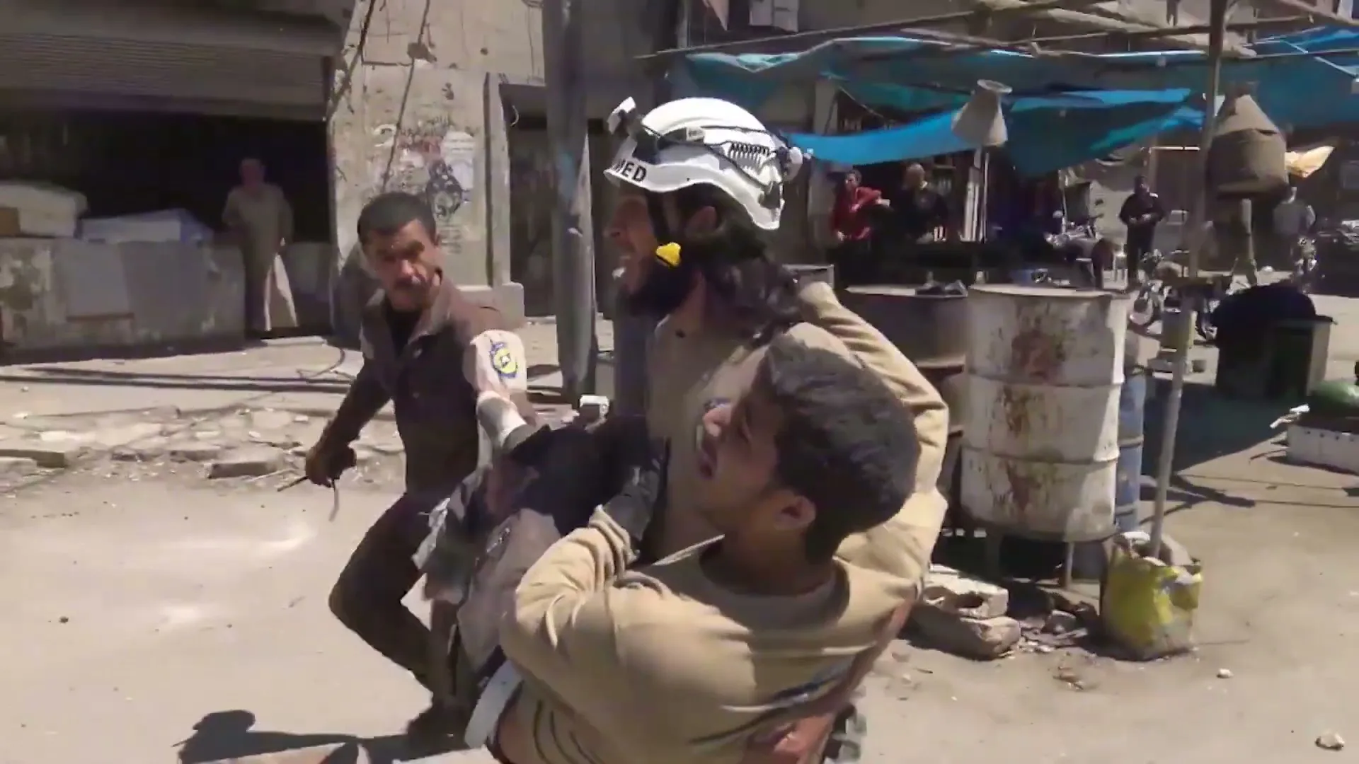 Two men carry an injured man through a rubble-strewn street in a war-torn city, with buildings and debris in the background.