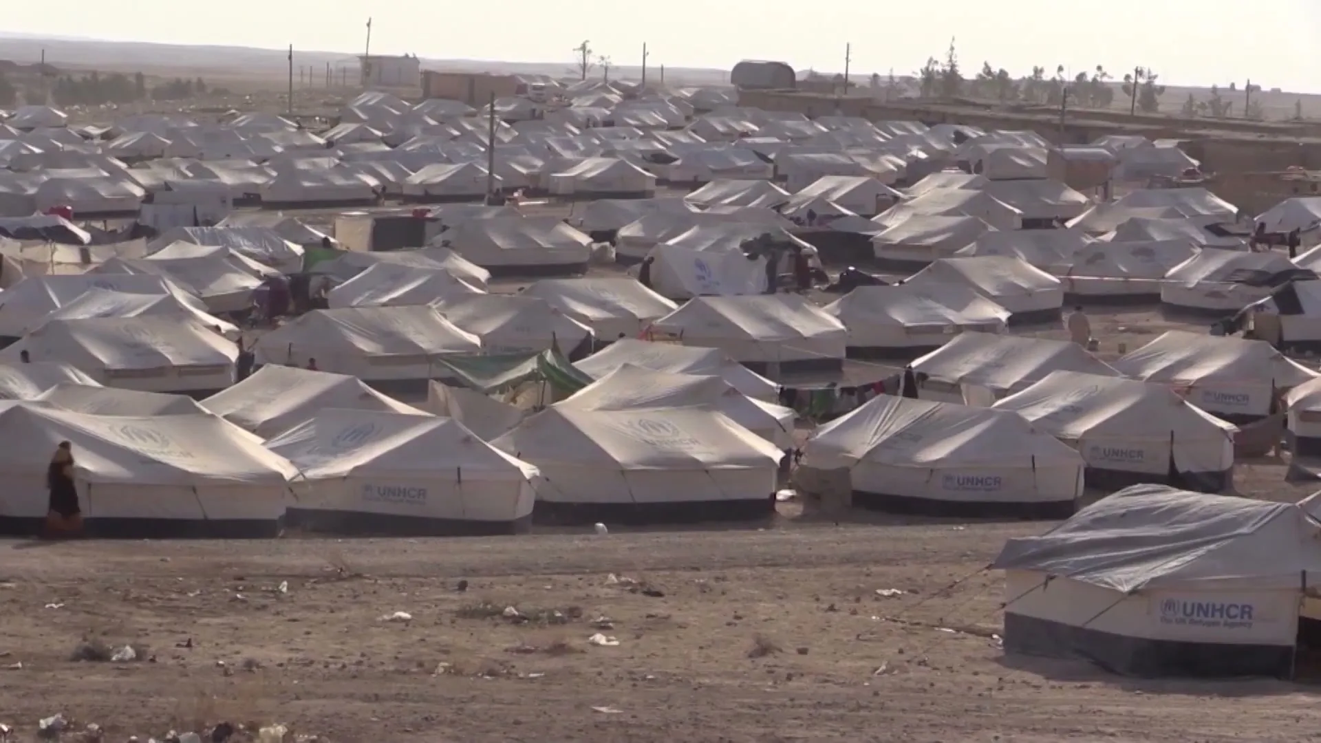 A wide shot shows a vast refugee camp filled with numerous white tents under a bright, hazy sky in a desert landscape.
