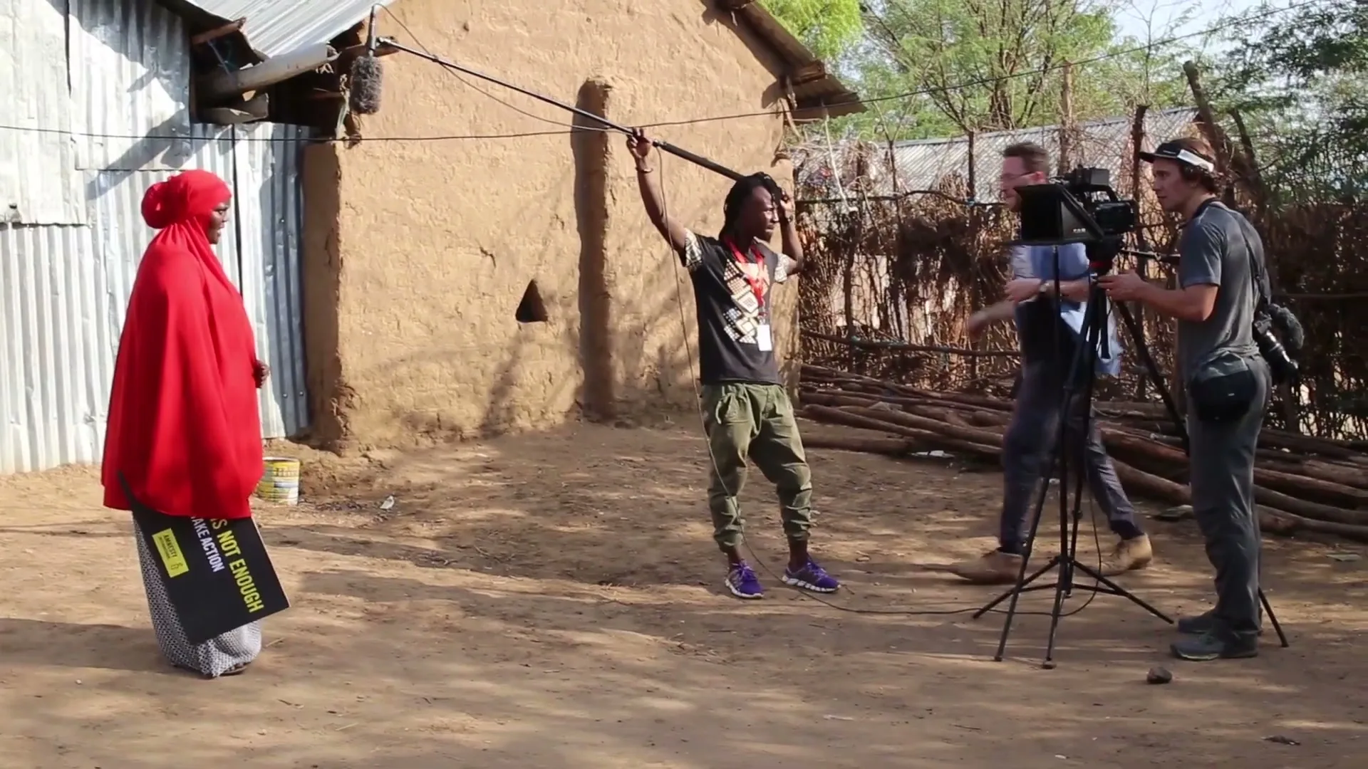 A woman in a red hijab holds a sign while a film crew records her in a rural village.