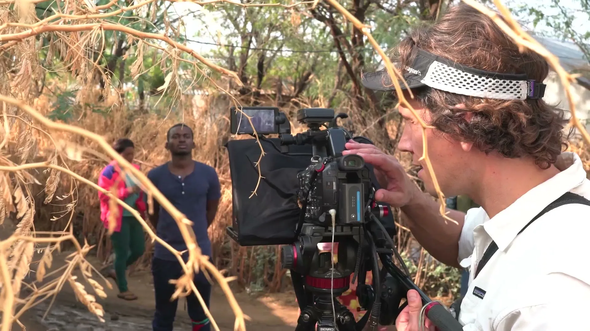 A male cameraman with curly hair and a visor operates a professional video camera, filming a young Black man standing in a rural, dusty outdoor setting.