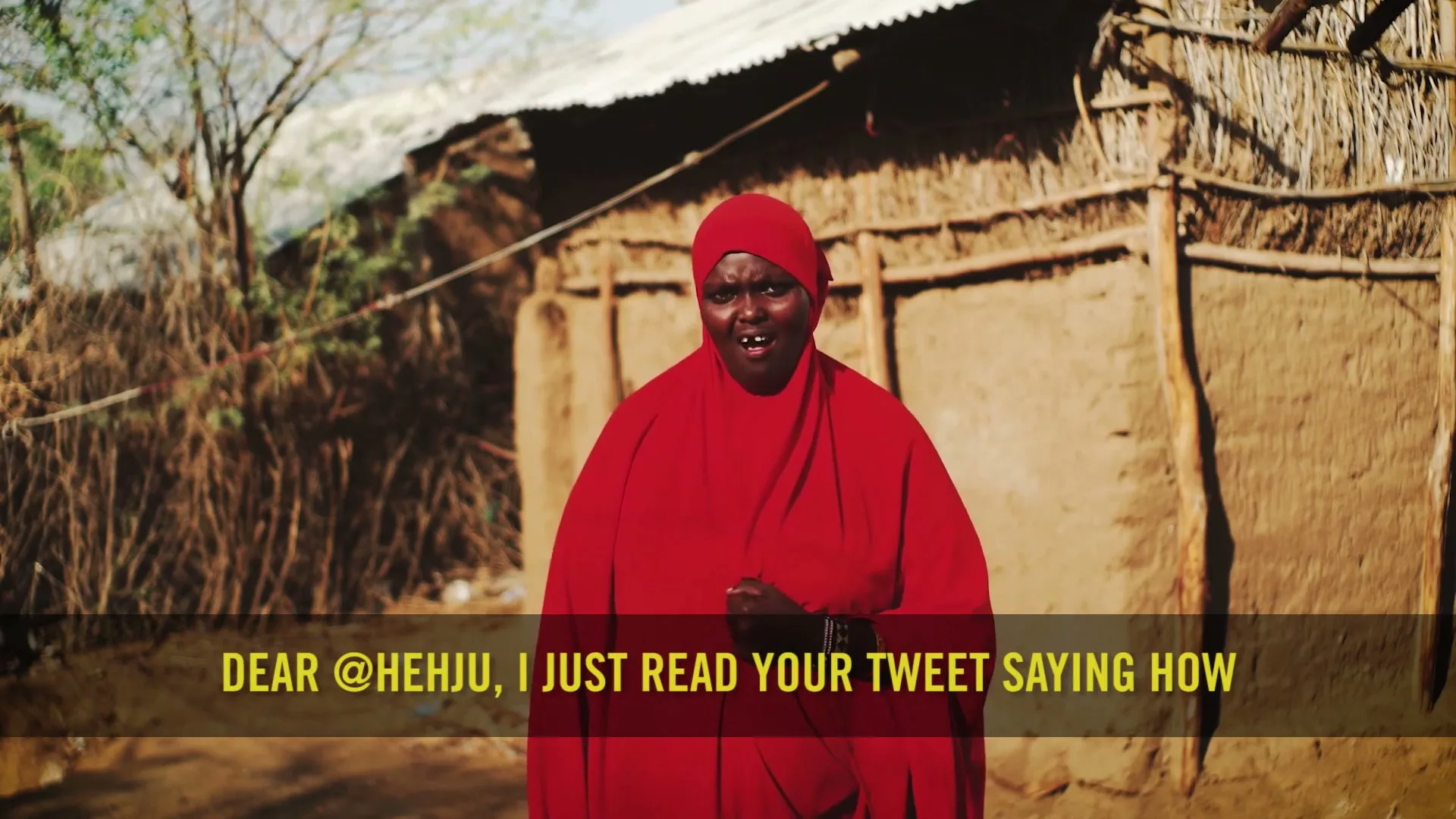 A woman in a red hijab and dress stands in front of a mud hut, speaking directly to the camera about the refugee crisis.