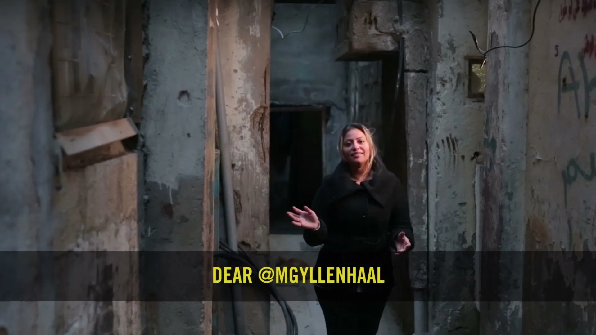 A woman in her 30s, wearing a black coat, stands in a narrow, dilapidated alleyway, speaking directly to the camera.