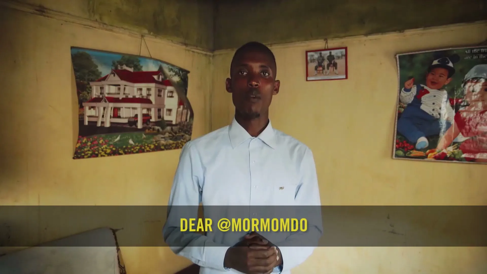 A young Black man in a light blue shirt stands in a room with yellow walls and pictures, speaking directly to the camera.